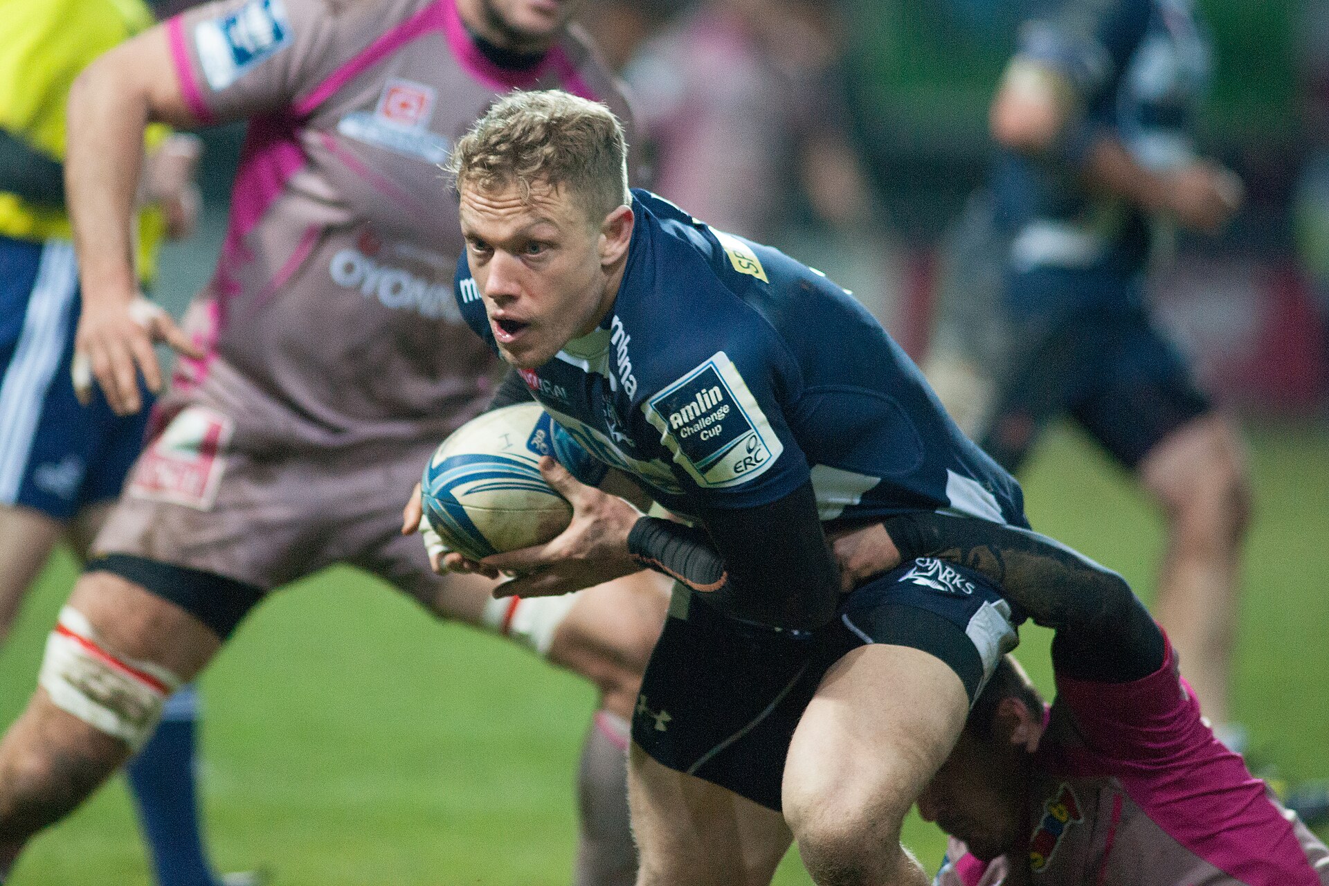 A rugby player in a blue jersey with 'Amlin Challenge Cup' on his sleeve runs with the ball while being tackled by an opponent in a pink jersey.