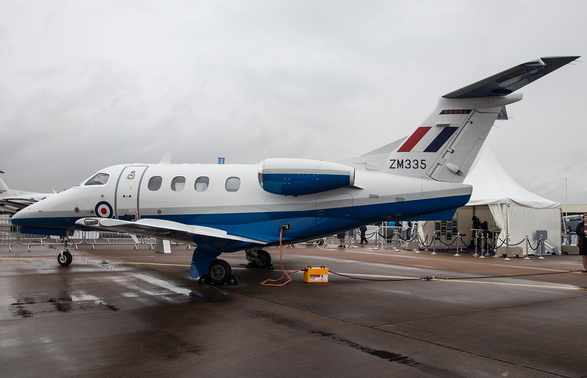 A white and blue jet aircraft with the registration ZM335 on its tail is parked on a wet tarmac. The aircraft has the Royal Air Force roundel on its side. In the background, there is a white tent and some people.
