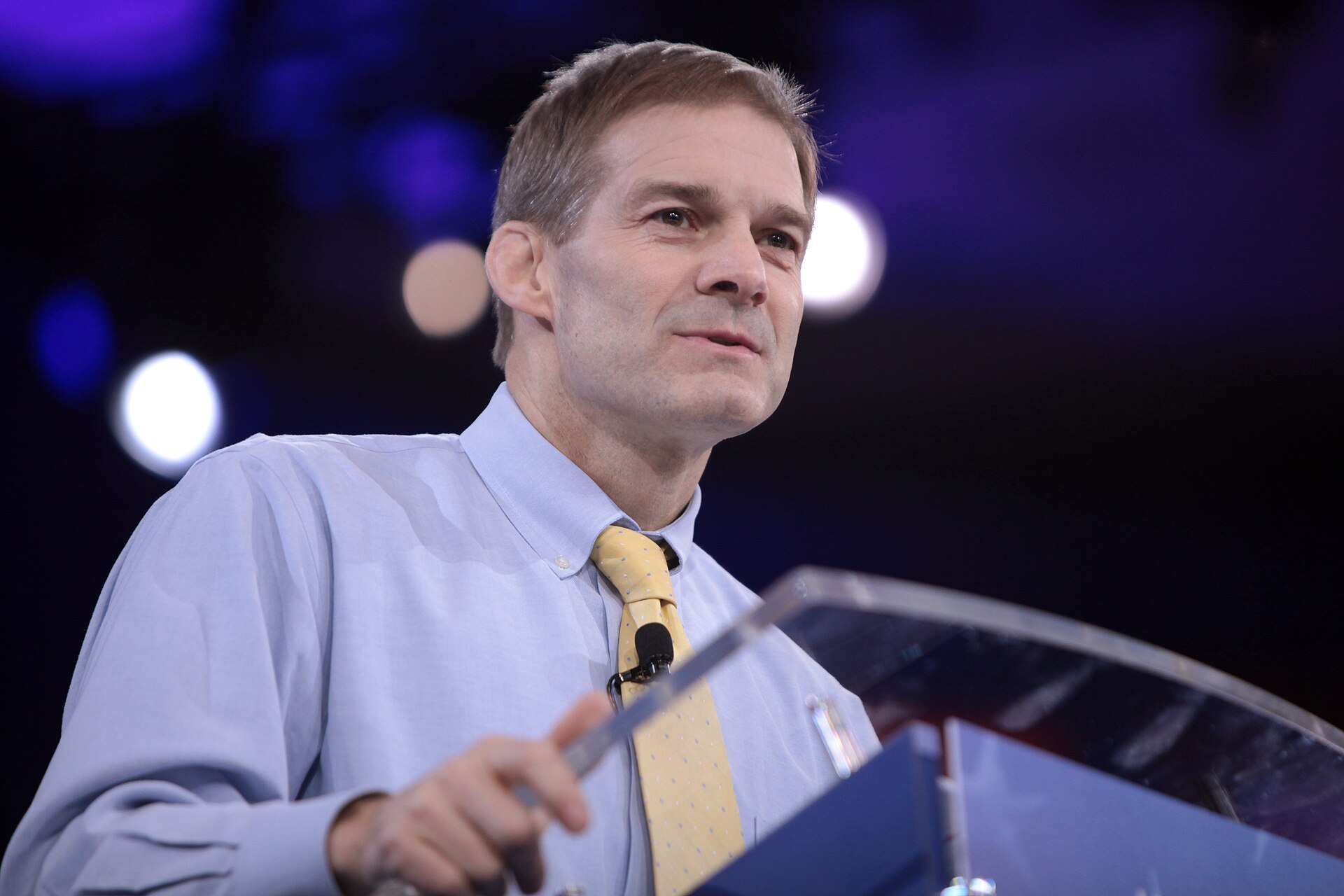 man in blue shirt and yellow tie speaking at a podium