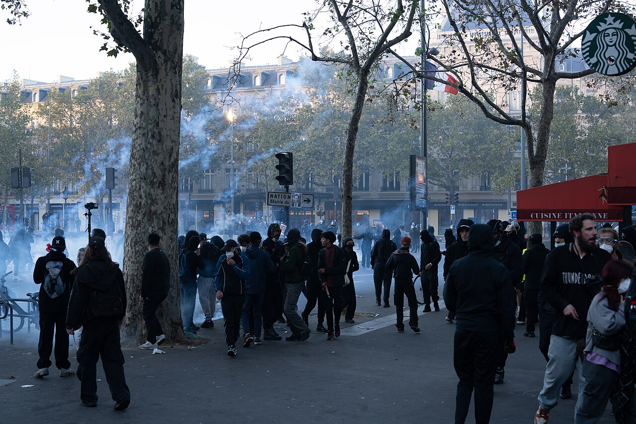 A crowd of people in black clothing are gathered on a street, with some holding up their hands. Smoke is visible in the background, and trees line the street. A Starbucks sign is visible on a building.