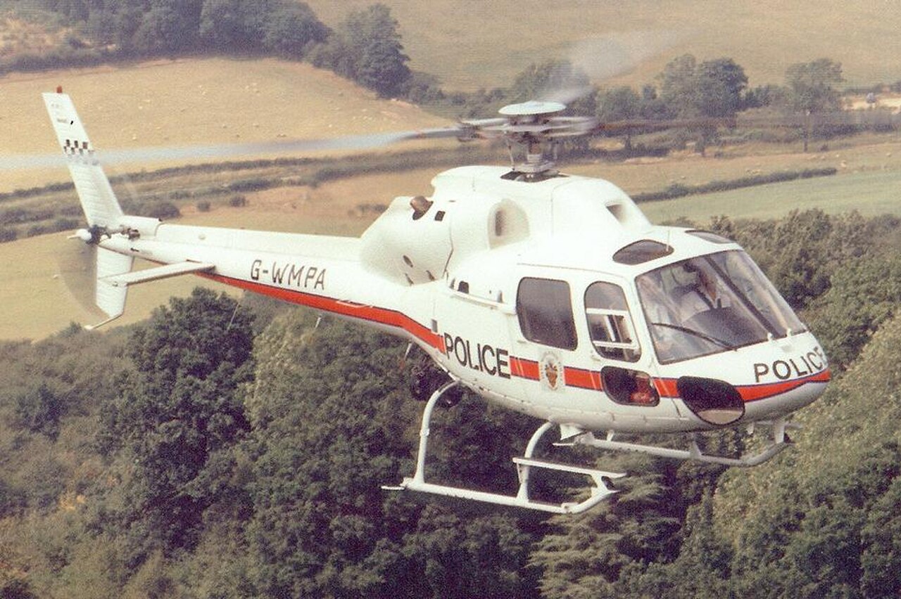 A white police helicopter with red stripes and the word POLICE written on its side, flying over a wooded area.