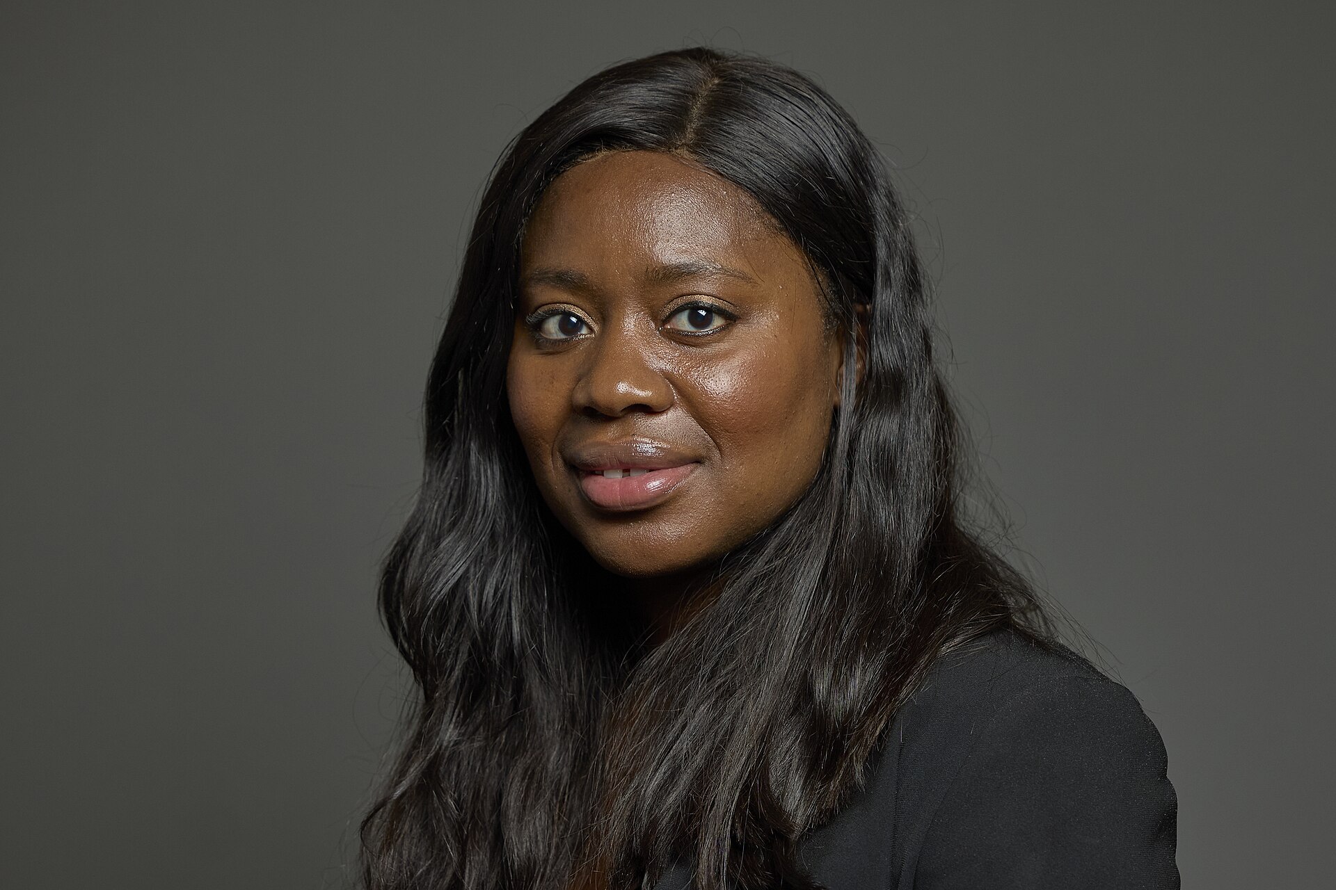 A headshot of Miatta Fahnbulleh, the Energy Secretary, looking towards the camera with a slight smile. She has dark skin, long dark wavy hair, and is wearing a black blazer.