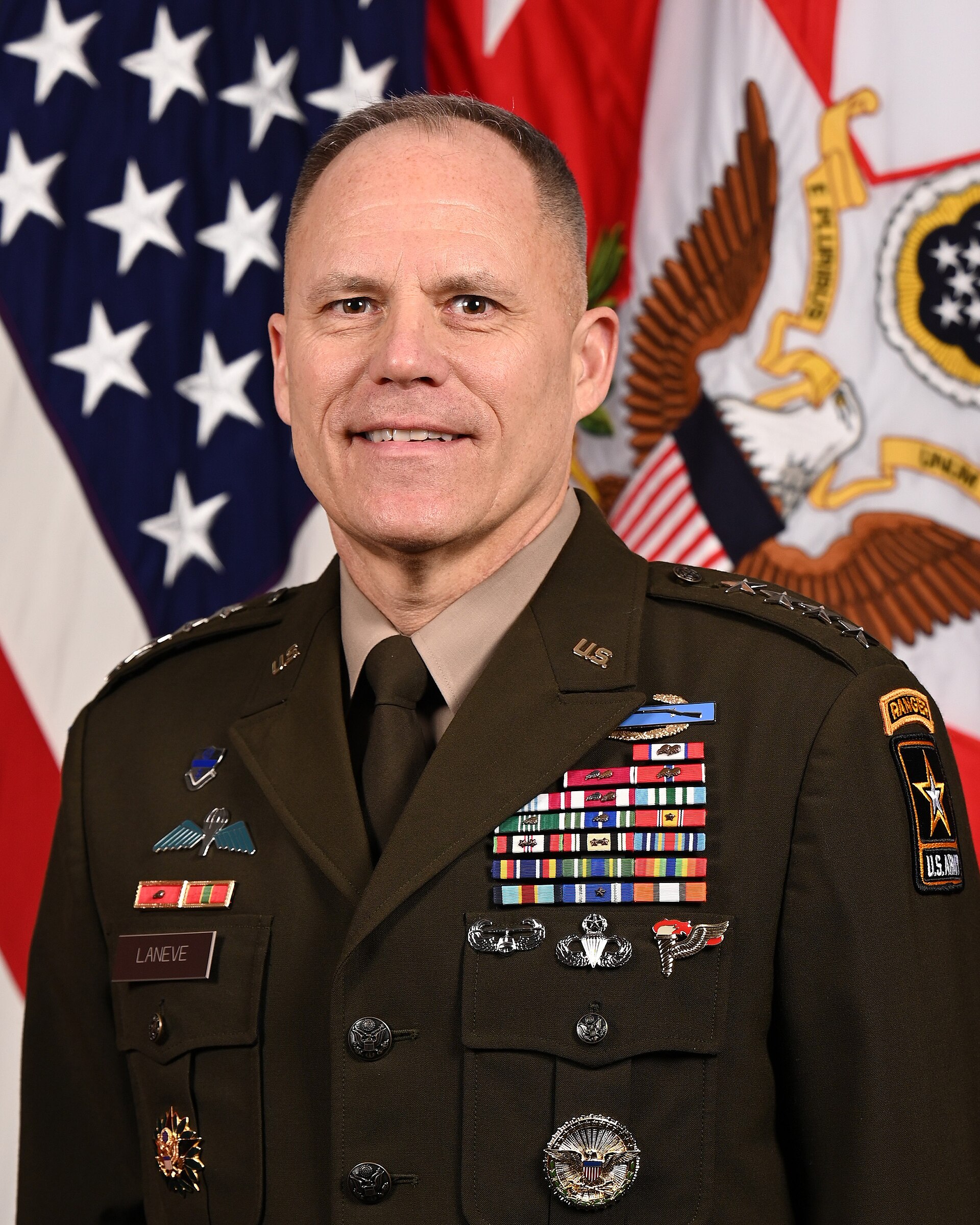 A portrait of a middle-aged man in a U.S. Army uniform adorned with numerous medals and insignia, standing in front of American and U.S. Army flags.