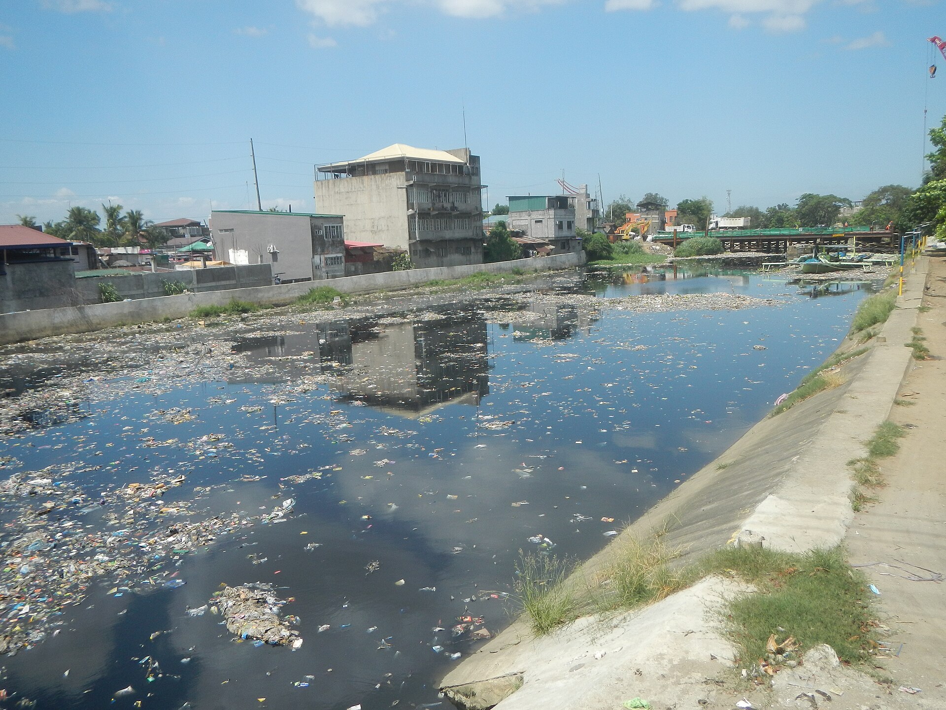 A river filled with trash and debris, with buildings and a bridge visible in the background under a clear blue sky.