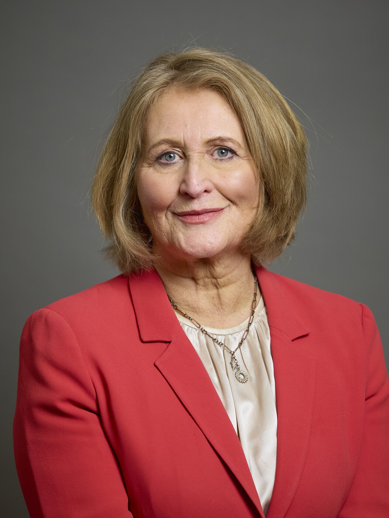 A middle-aged woman with short blonde hair, wearing a red blazer and a white blouse, smiling slightly at the camera.