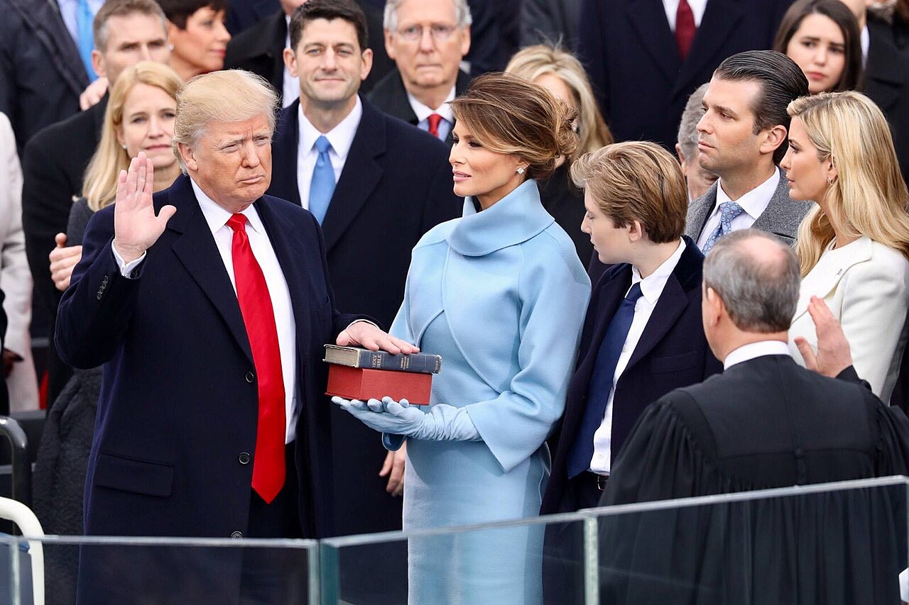 Donald Trump raises his hand while placing his other hand on a Bible, with Melania Trump and others standing behind him.