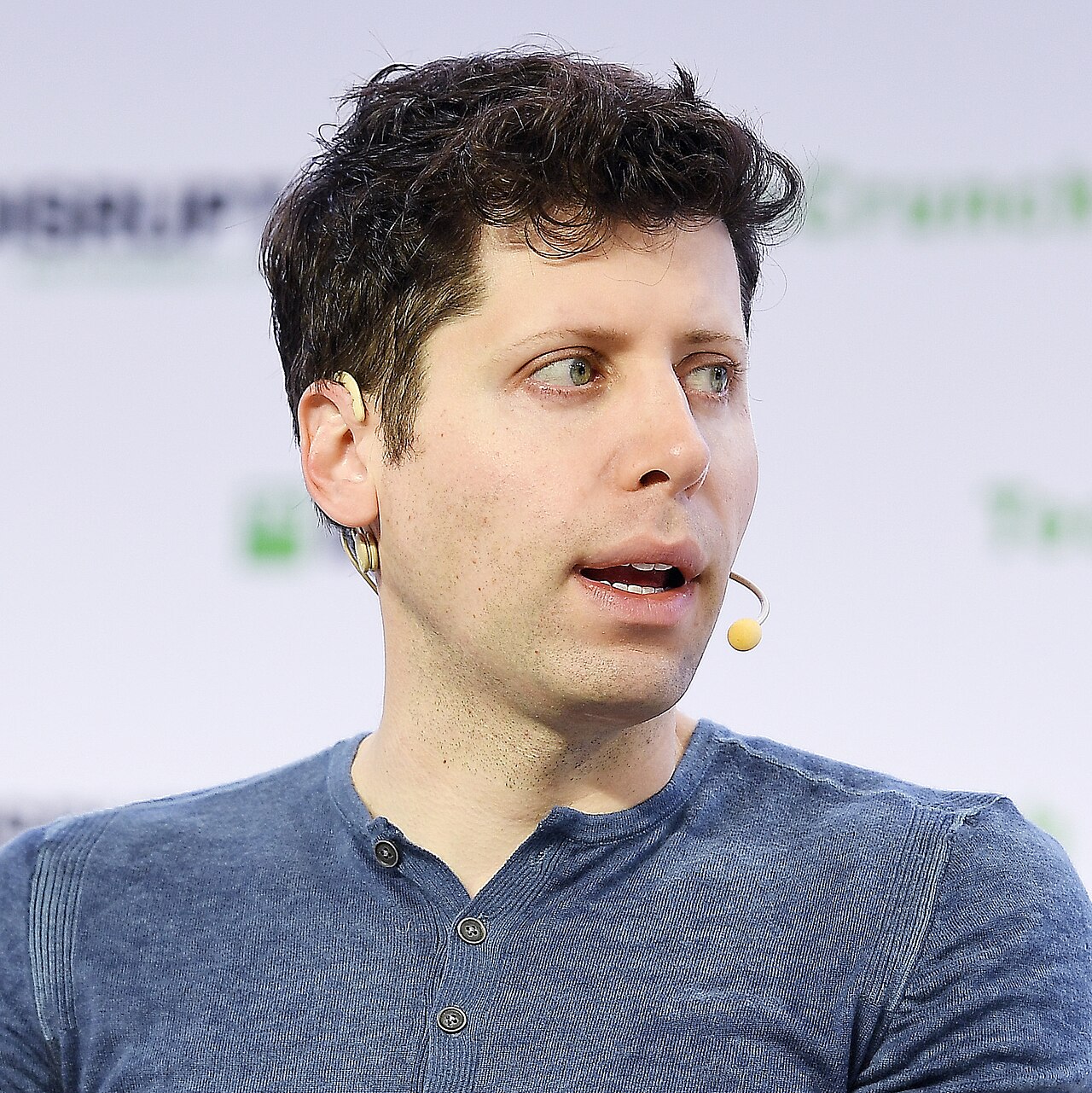 A man with short dark hair and a blue shirt, wearing a small earpiece with a yellow ball on the end, looking to the right with a slight smile.