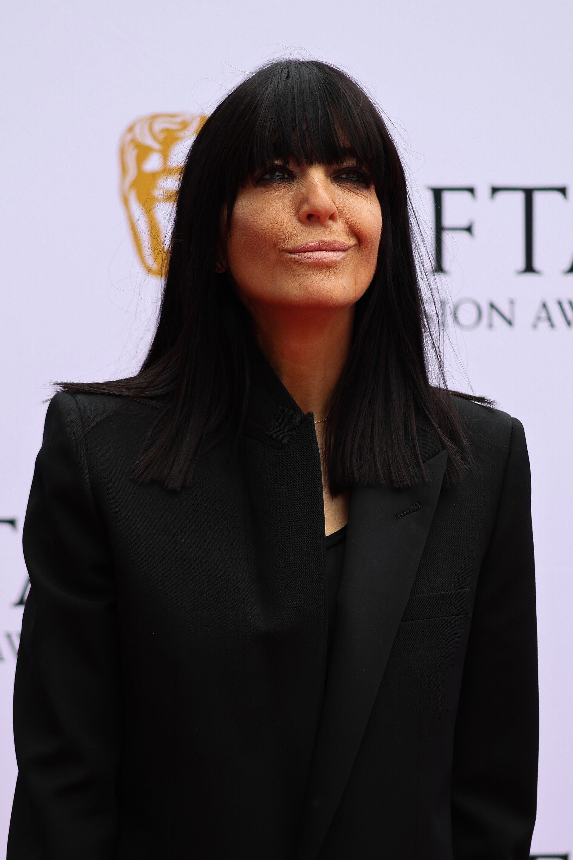 Claudia Winkleman, a middle-aged woman with long dark hair and bangs, wearing a black blazer and top, standing in front of a white backdrop with a gold and black logo.