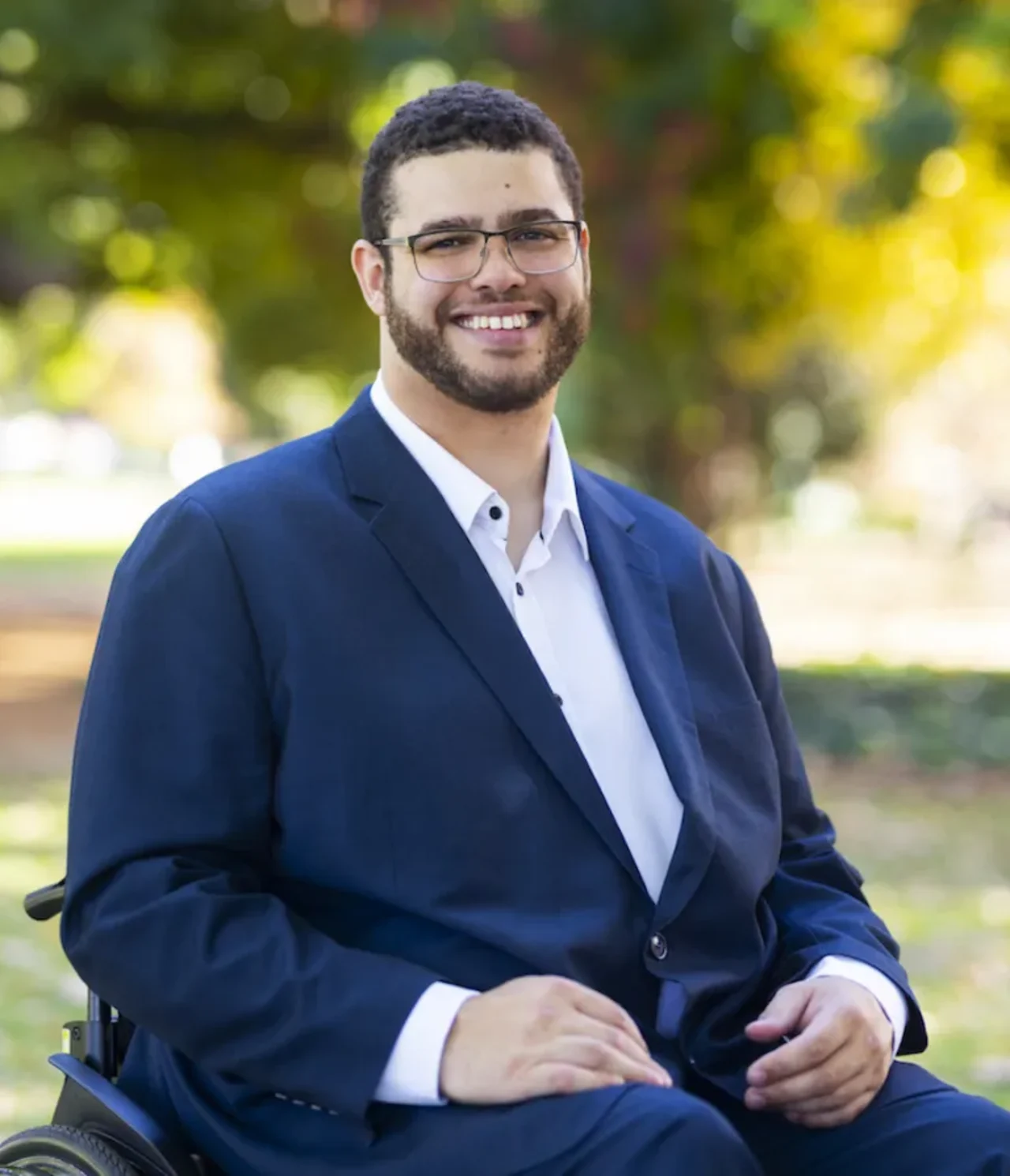 A man in a wheelchair wearing a dark blue suit and white shirt smiles at the camera, with a blurred background of trees and sunlight.