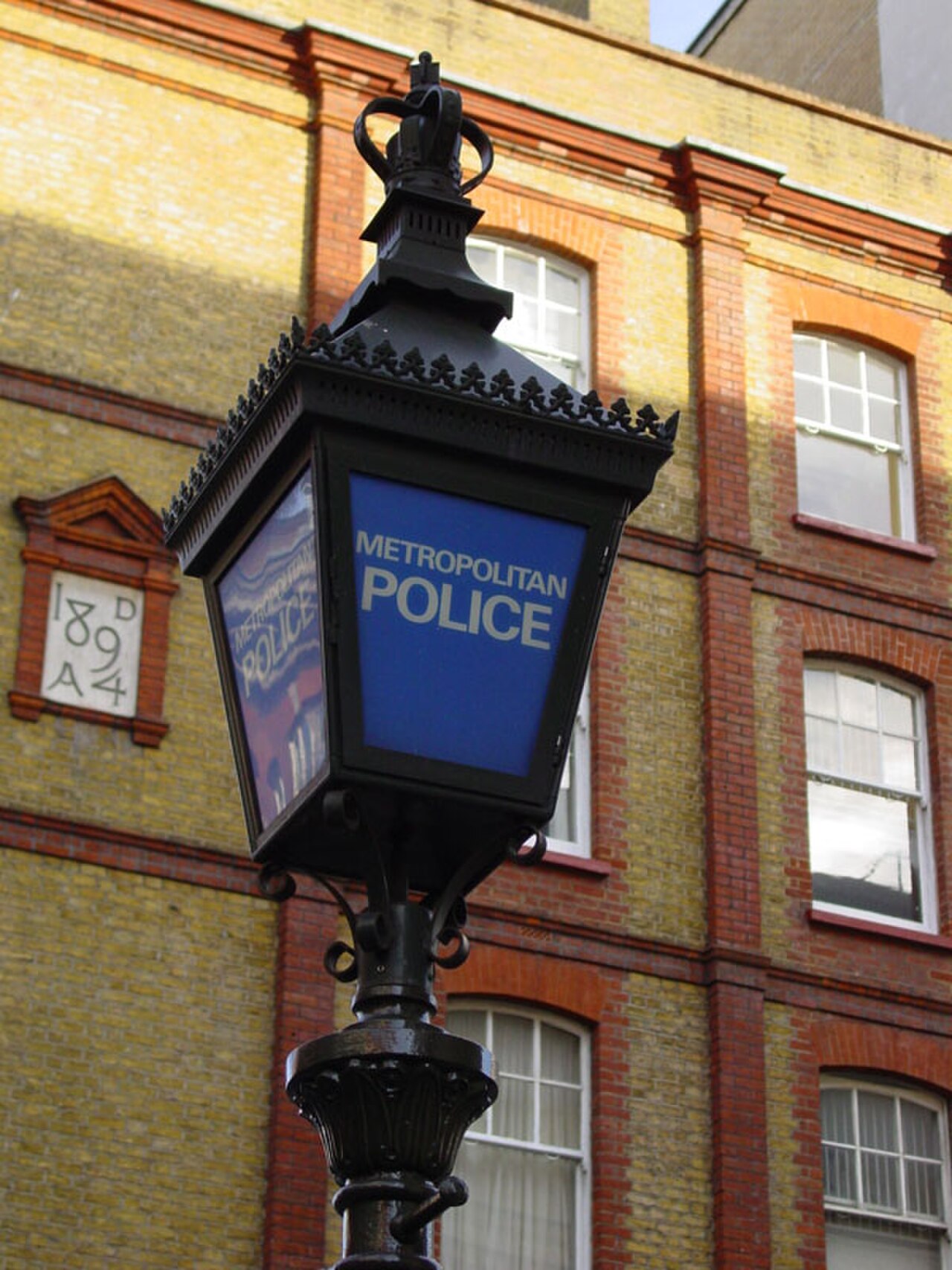 A black ornate lamppost with a blue sign that reads "METROPOLITAN POLICE" is in front of a yellow brick building with red brick accents and white-paned windows.