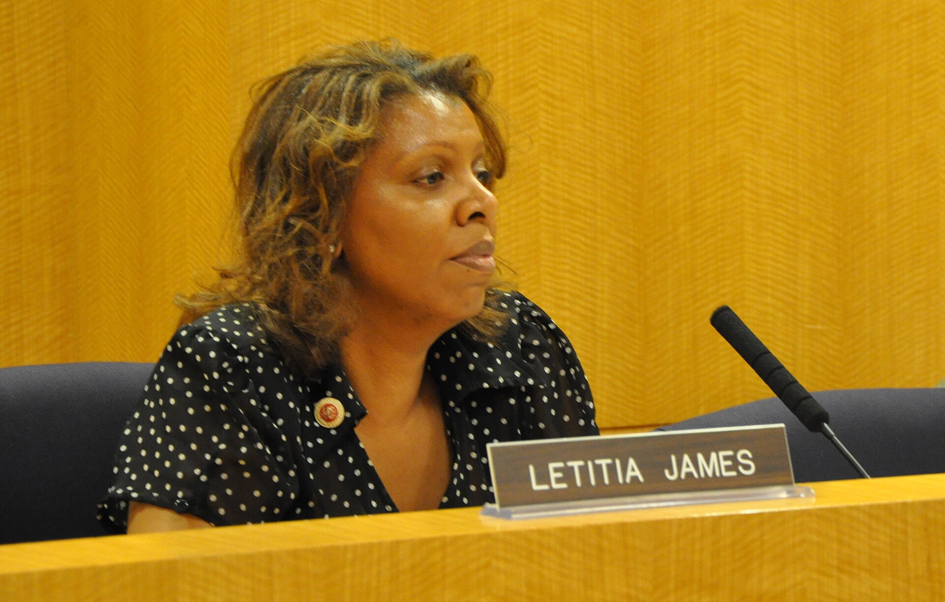 A woman with shoulder-length brown hair wearing a black blouse with white polka dots and a gold brooch sits at a desk with a nameplate that reads LETITIA JAMES.