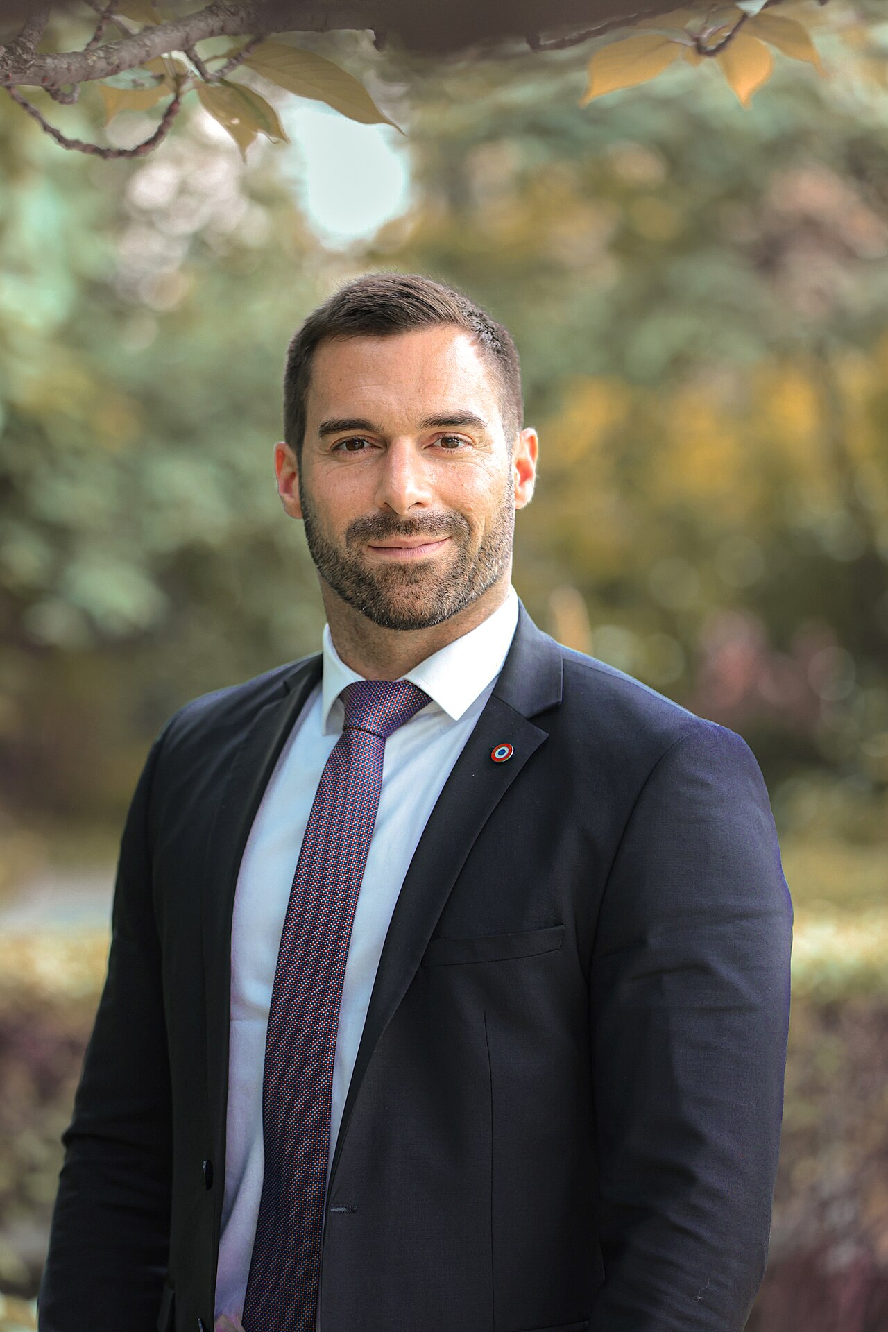 A smiling man in a suit and tie standing in front of a blurred background of trees and foliage.