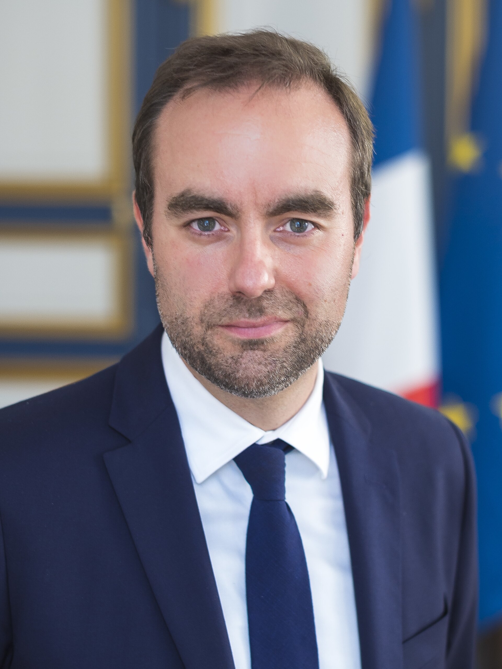 A man in a blue suit and tie stands in front of a French flag.
