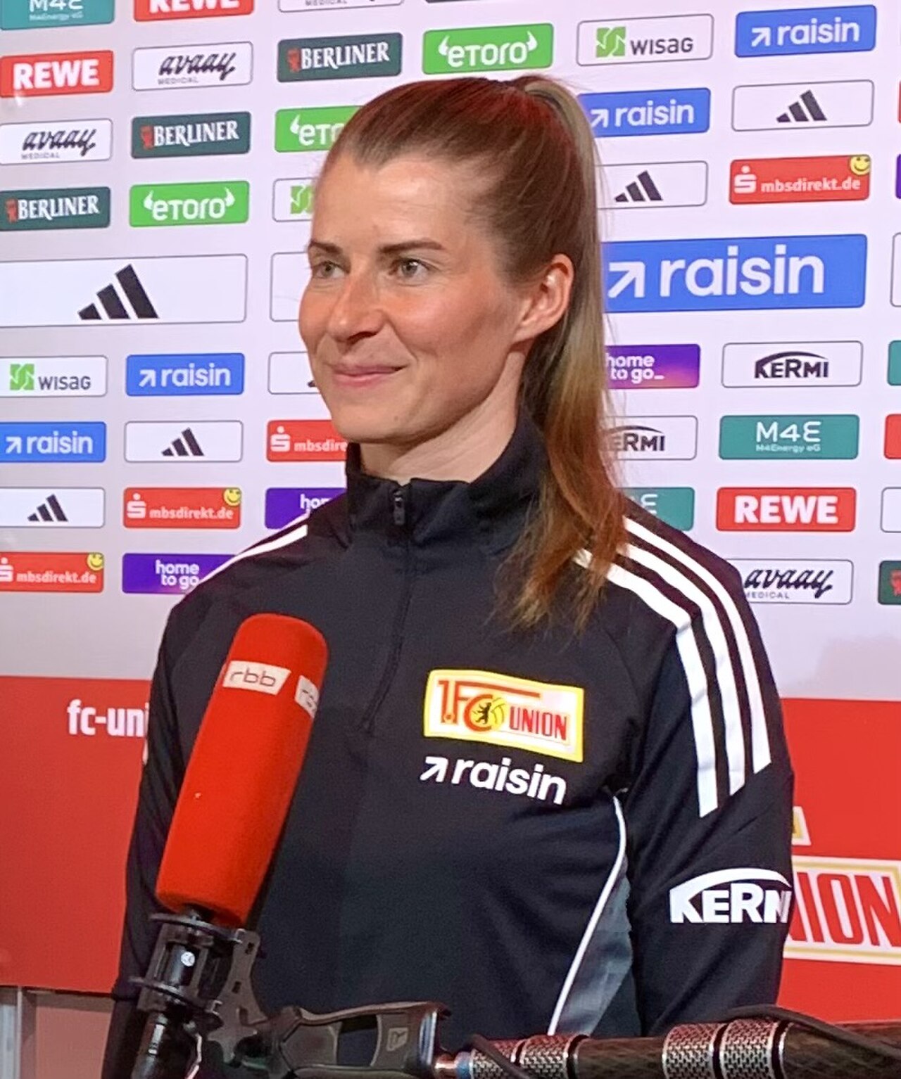 A middle-aged woman with long brown hair pulled back into a ponytail, wearing a black tracksuit with white stripes and logos, stands in front of a wall of sponsor logos and speaks into a red microphone.