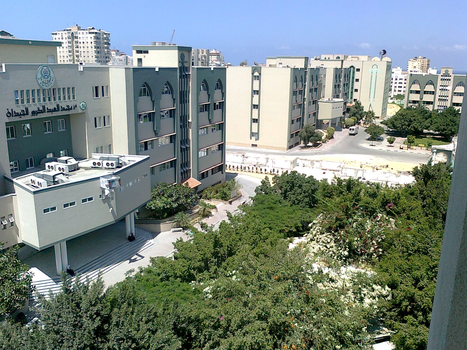 A wide shot of the Islamic University of Gaza campus, showing multiple modern buildings with green and beige facades, surrounded by trees and clear blue sky.