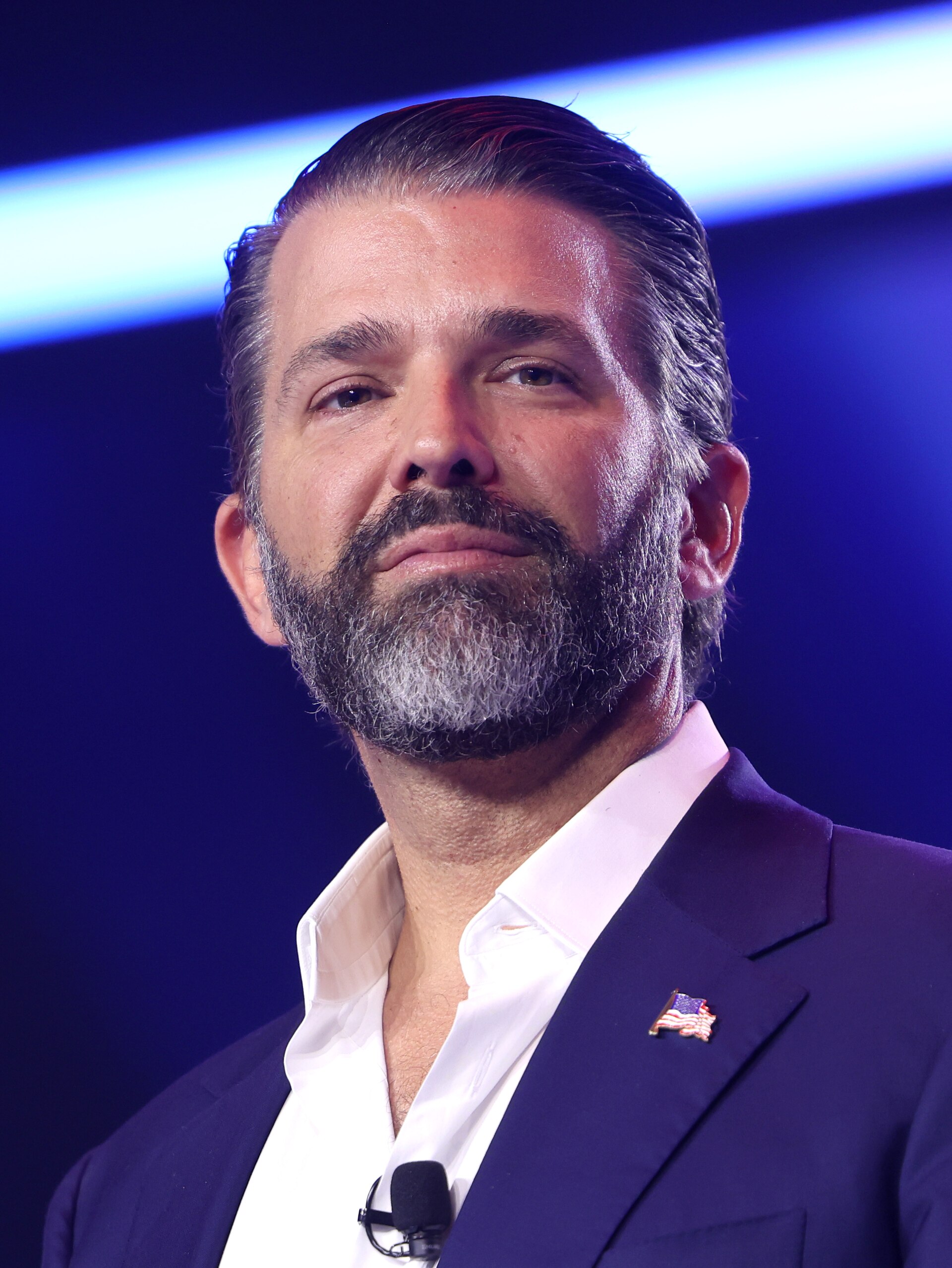 Donald Trump Jr. wearing a navy suit jacket, white shirt, and American flag pin, with a microphone visible.