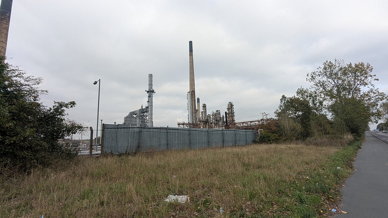An industrial facility with multiple smokestacks and pipes, surrounded by a fence and situated near a road and grassy area.