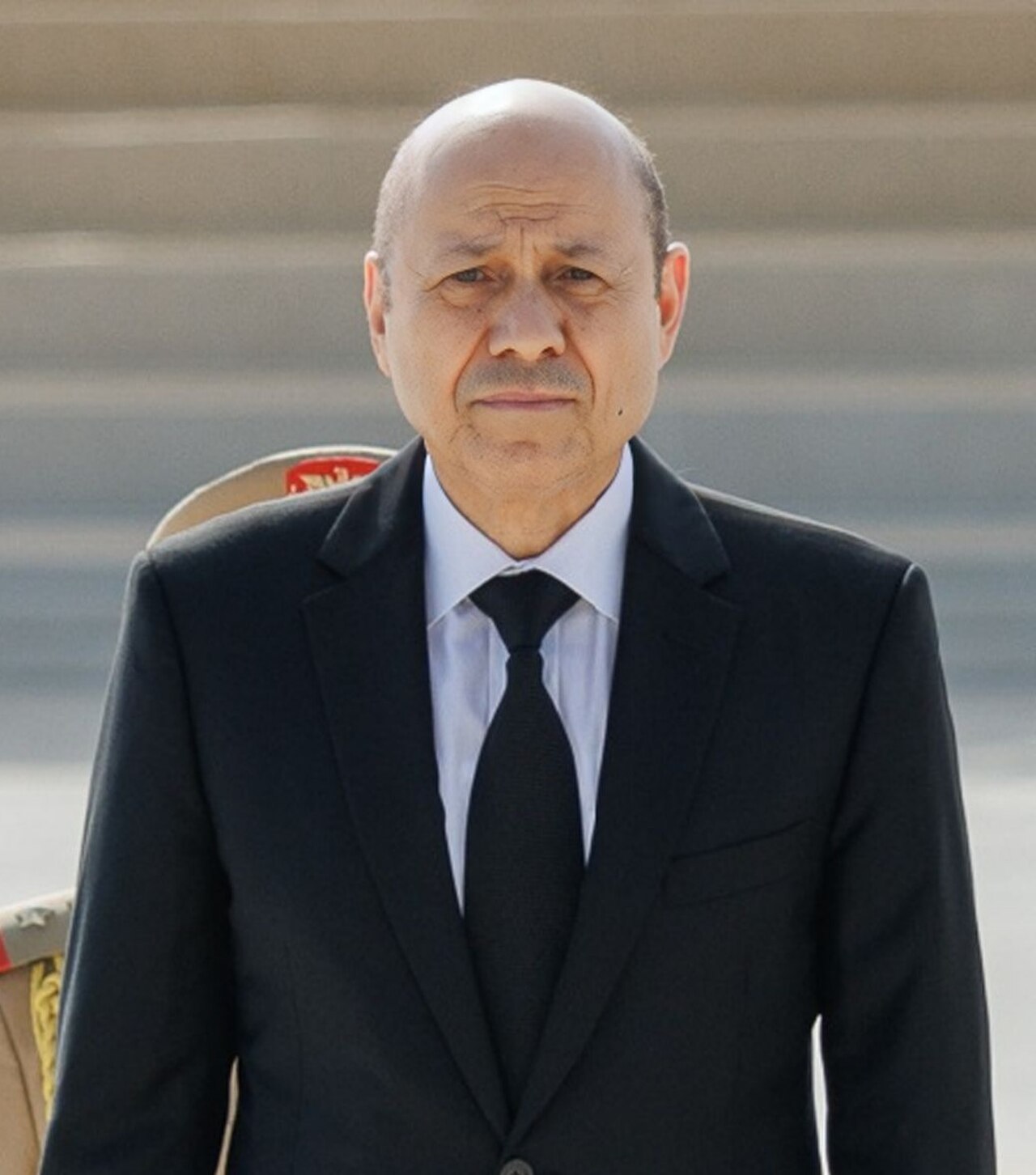 A man in a black suit, white shirt, and black tie stands facing the camera.