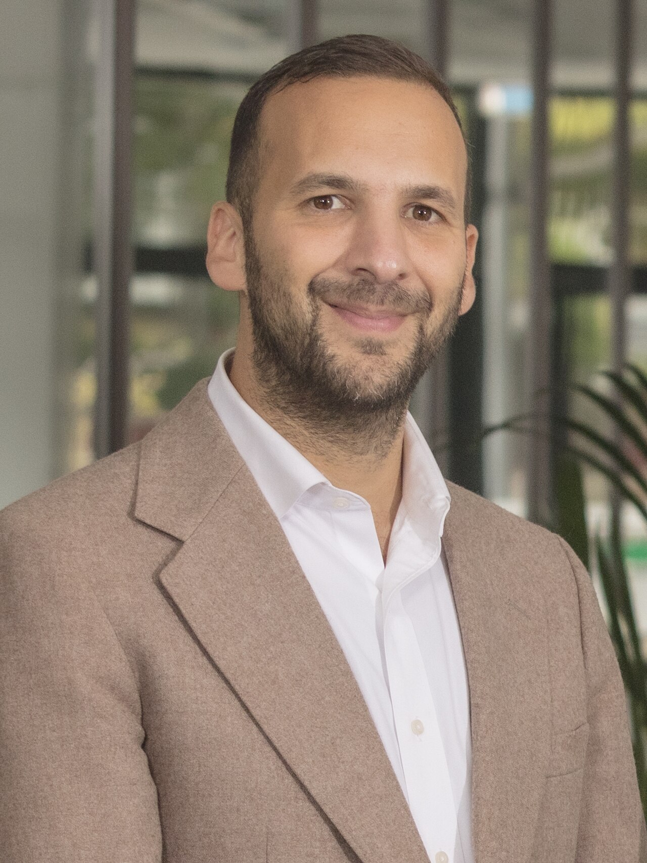 A man with short dark hair and a beard, wearing a white collared shirt and a light brown blazer, smiles at the camera.