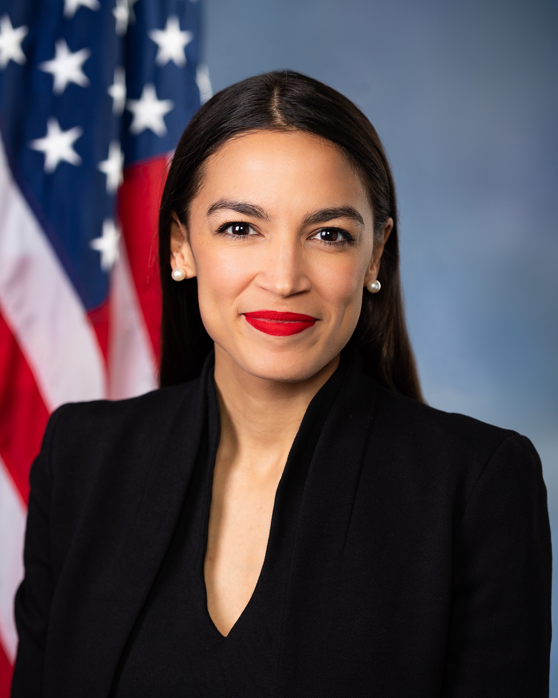 Alexandria Ocasio-Cortez smiles in front of an American flag.