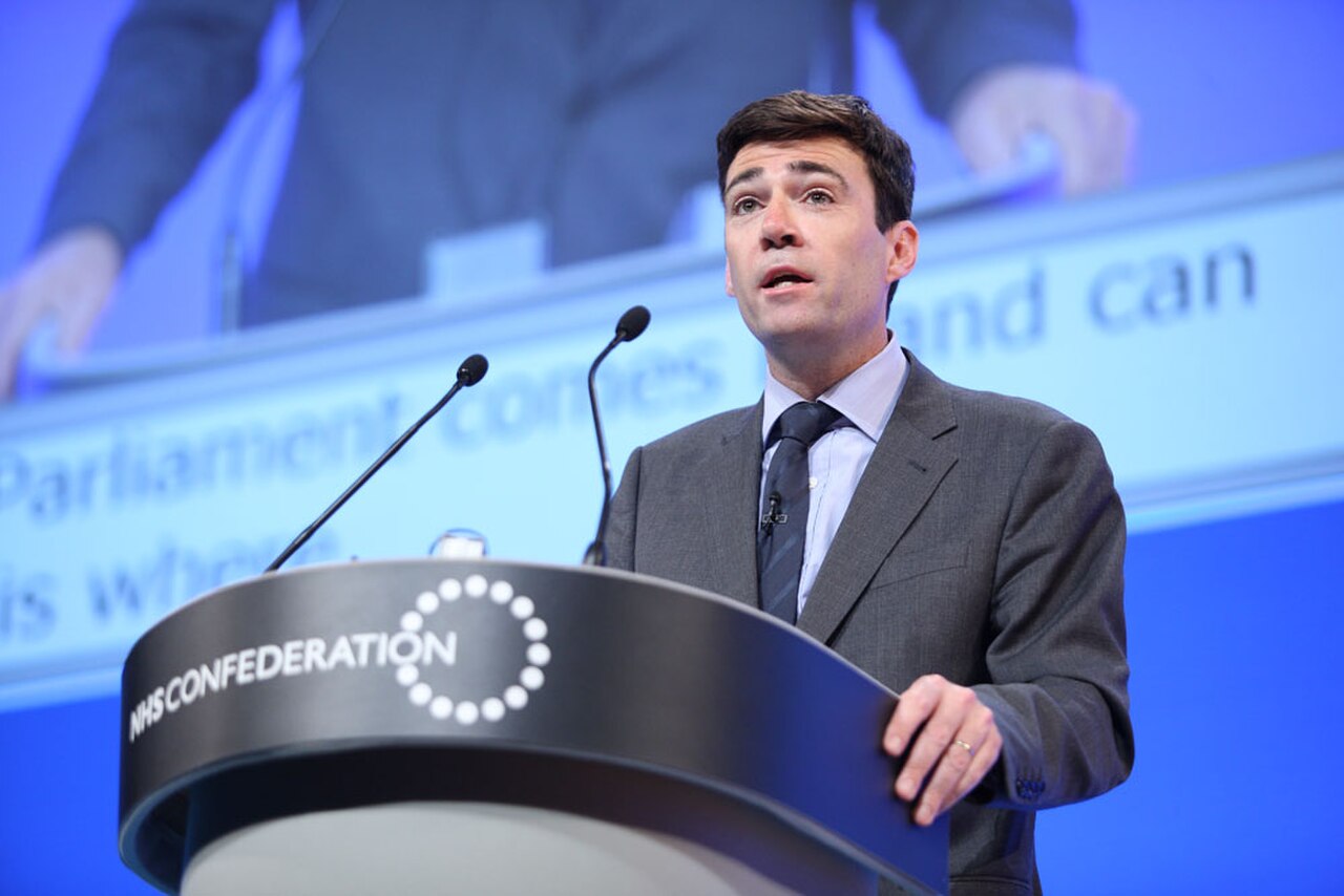 A man in a gray suit speaking at a podium with two microphones, the podium has a NHS Confederation sign on the front.