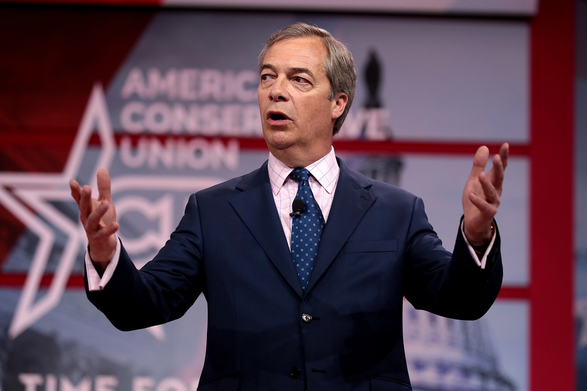 Nigel Farage speaking at a podium with his hands gesturing outwards. Behind him is a banner that reads "AMERICA CONSER UNION".
