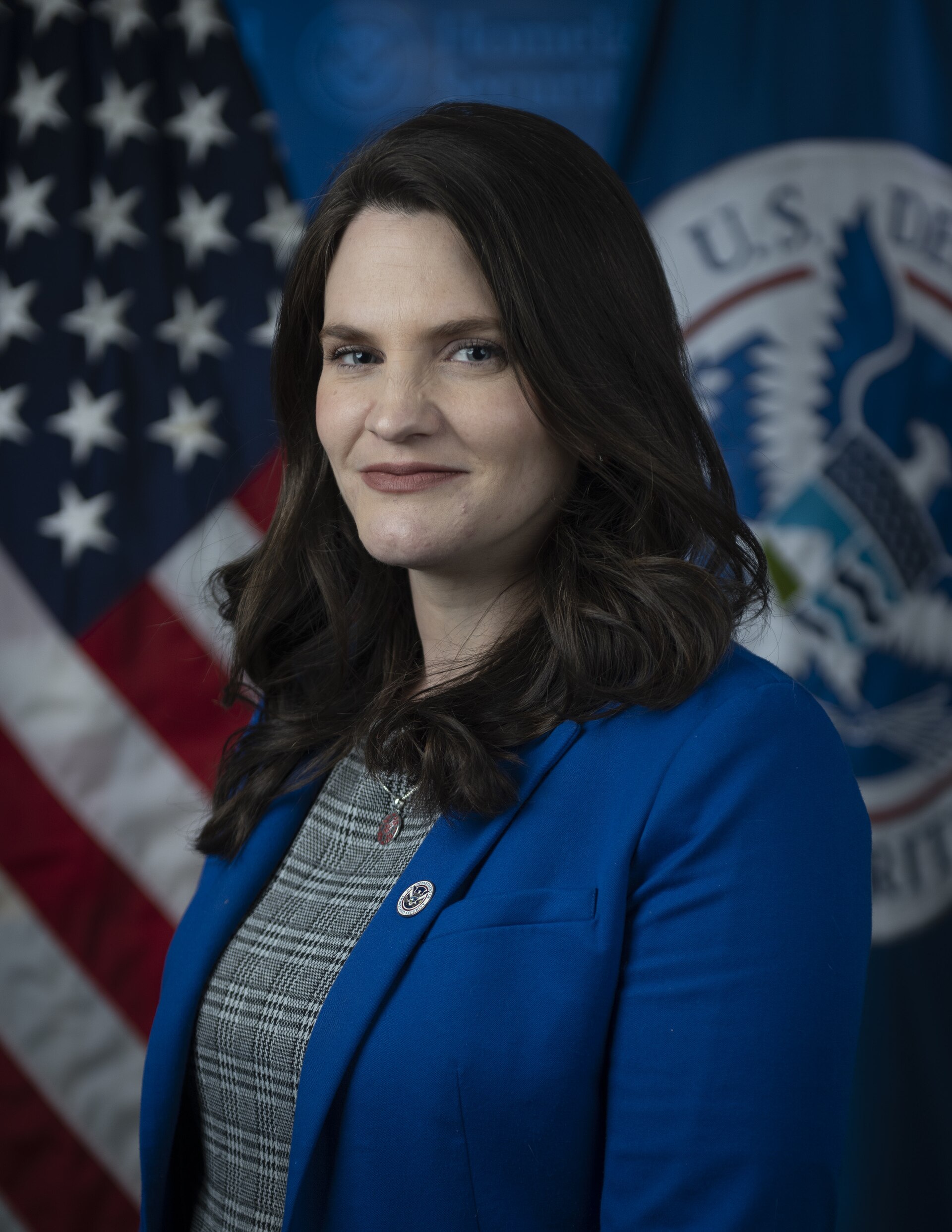 A woman with long brown hair wearing a blue blazer and gray shirt stands in front of an American flag and a U.S. Department of State flag.