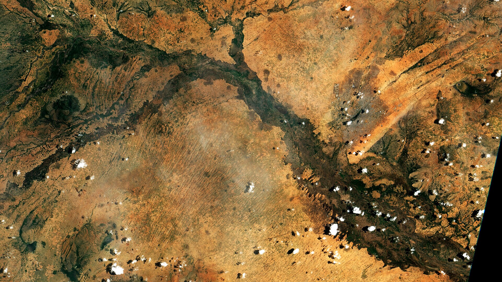 Satellite view of a dry, arid landscape with a winding river or dry riverbed cutting through it. Patches of green vegetation are visible along the riverbanks, contrasting with the surrounding brown and orange terrain. Scattered clouds dot the sky, casting shadows on the land below. Several small set