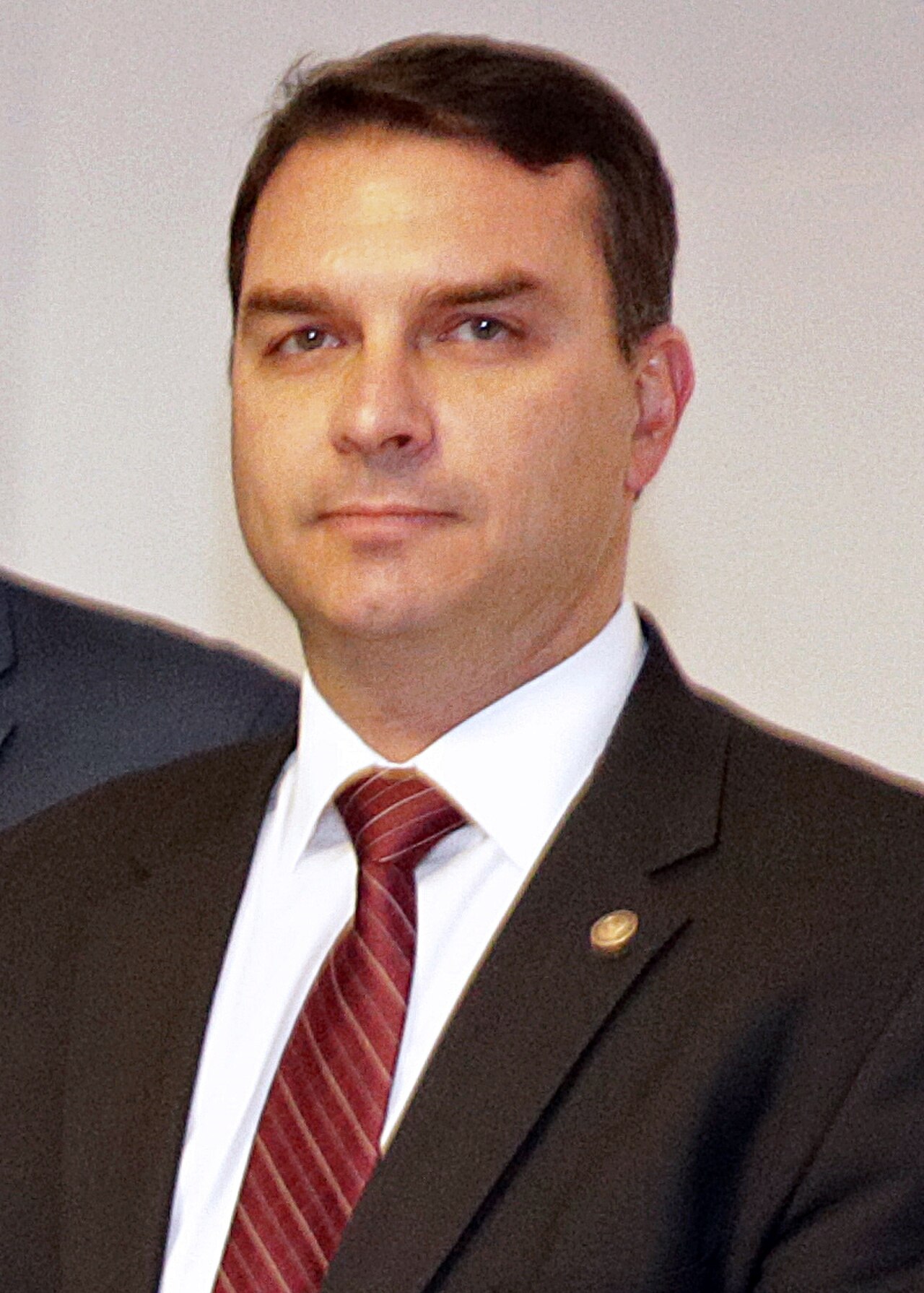 Flávio Bolsonaro, wearing a black suit, white shirt, and red tie, looking to the left with a neutral expression.