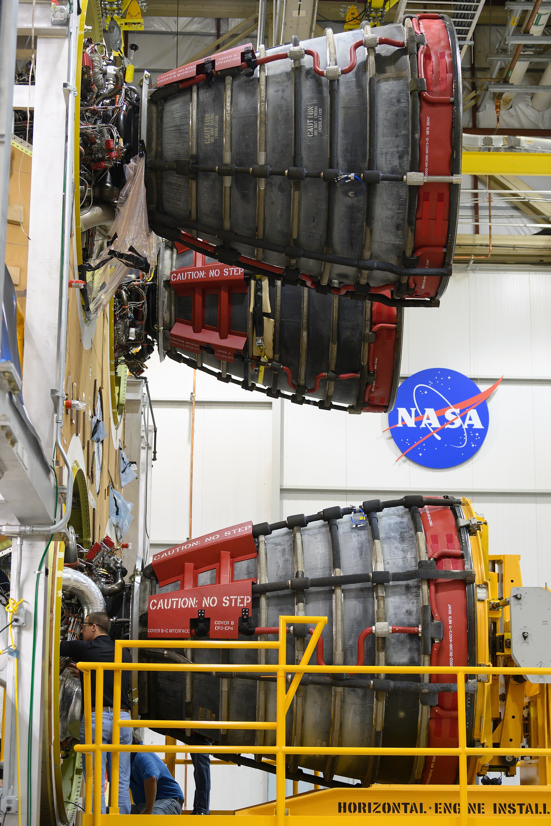 Two large rocket engines are mounted vertically in a hangar, with the NASA logo visible on the wall behind them. Technicians in blue jeans and shirts are working on the engines, which are surrounded by yellow scaffolding and safety railings.