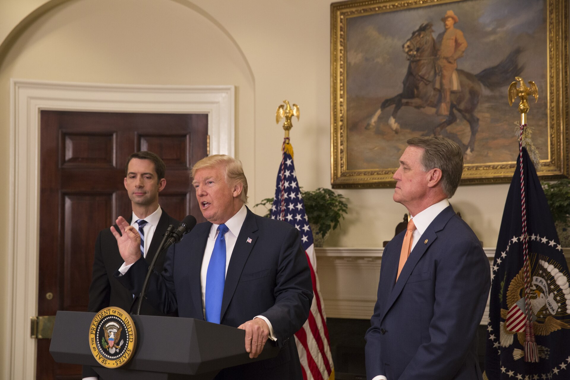 Donald Trump at a podium with two men beside him, speaking, with the Seal of the President of the United States on the podium, and flags and a painting in the background.