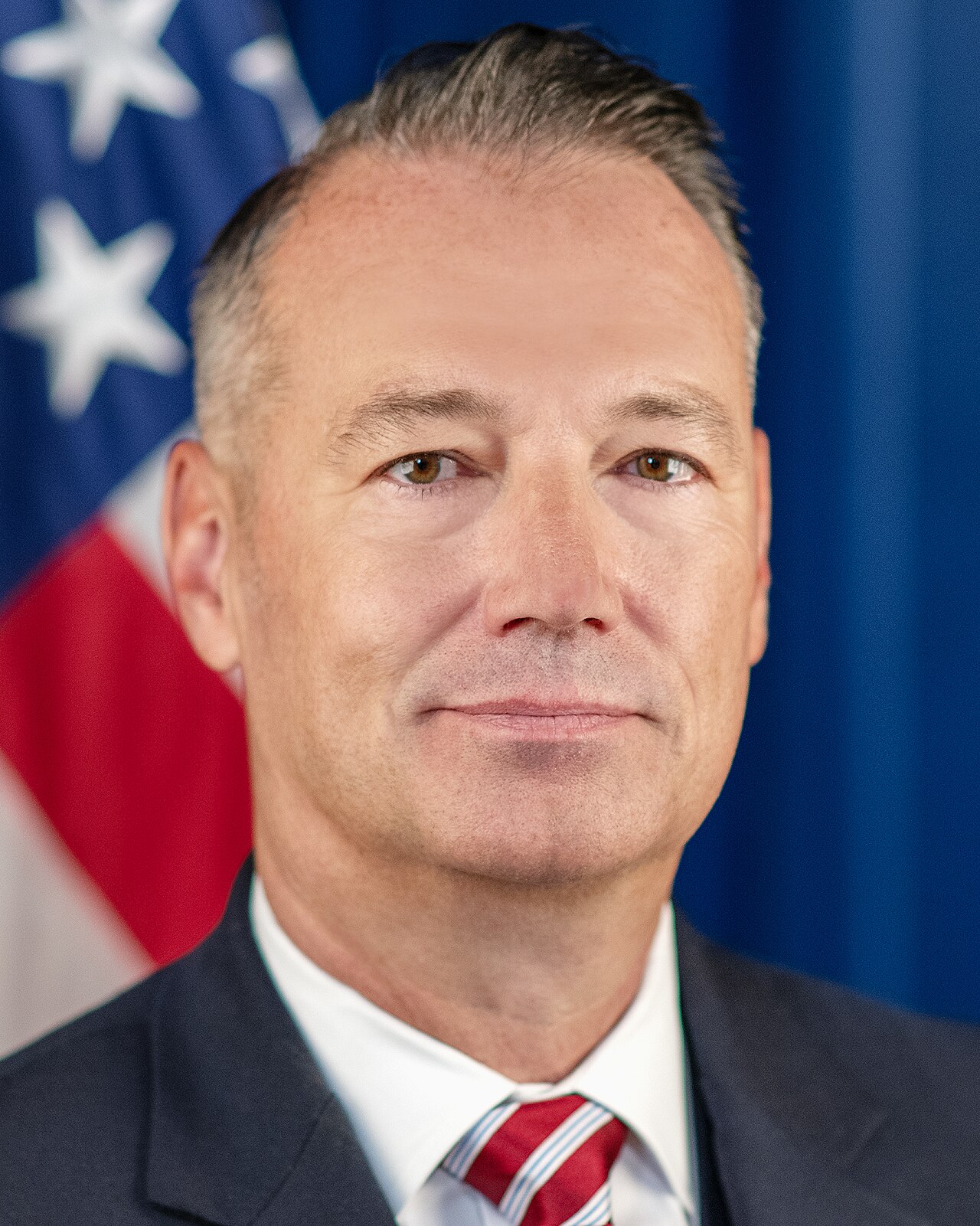 A middle-aged man with short graying hair, wearing a suit and tie, in front of an American flag.