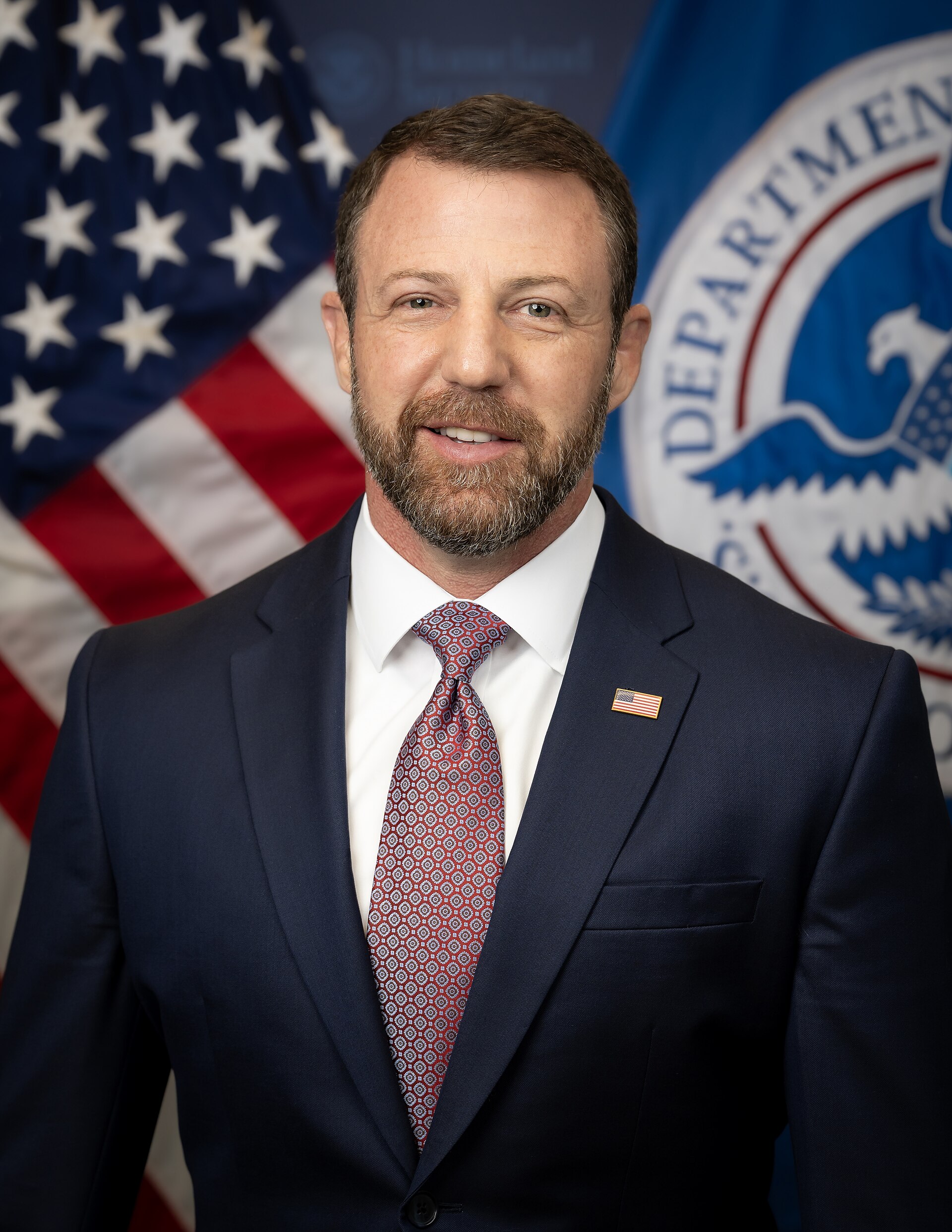 A man in a suit standing in front of an American flag and a Department of Homeland Security logo.