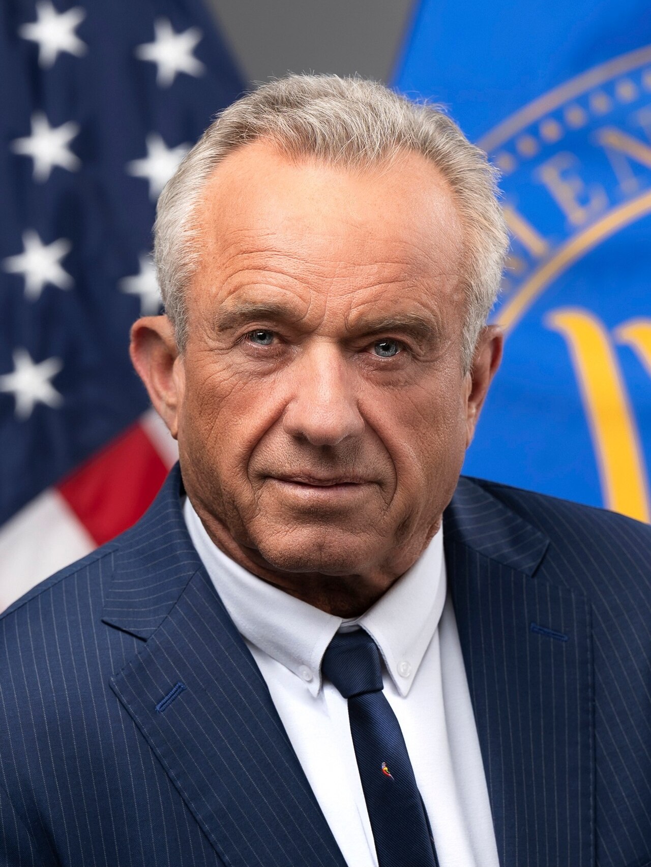 A man with short gray hair wearing a blue suit and tie stands in front of an American flag and another flag.
