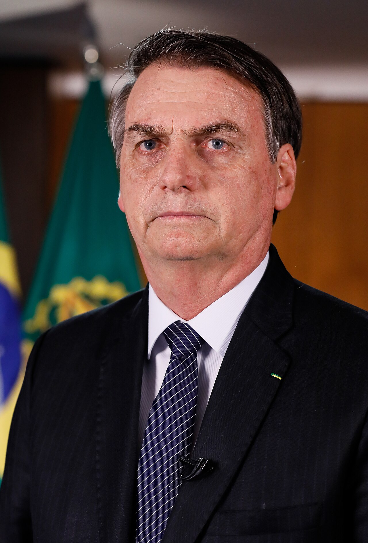 A man in a black suit, white shirt, and blue tie with diagonal white stripes, with a small green pin on his left lapel, stands in front of a blurred background featuring a green flag with a yellow emblem.