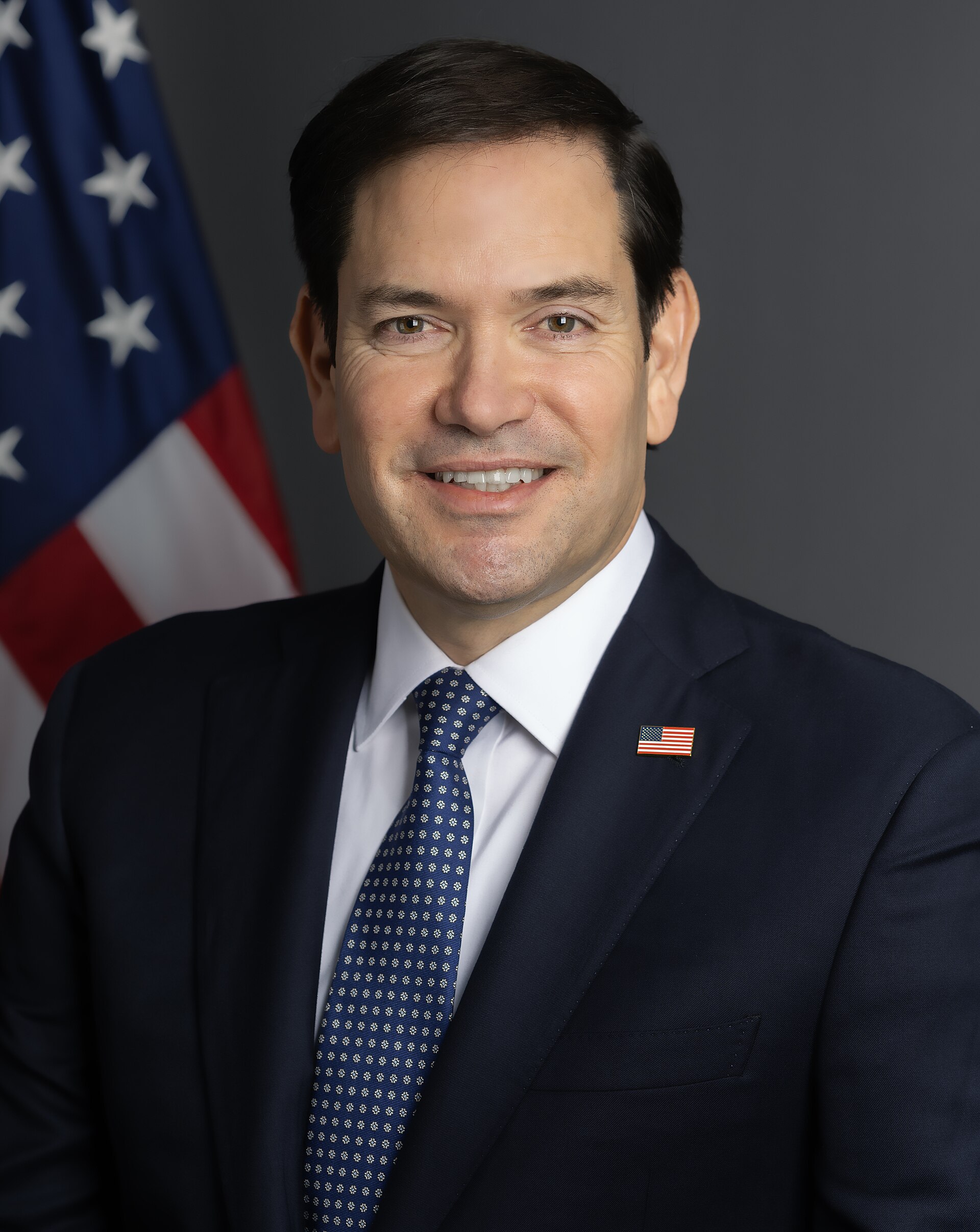 Marco Rubio, a middle-aged man with dark hair, wearing a navy suit, white shirt, blue tie, and American flag lapel pin, stands in front of an American flag.