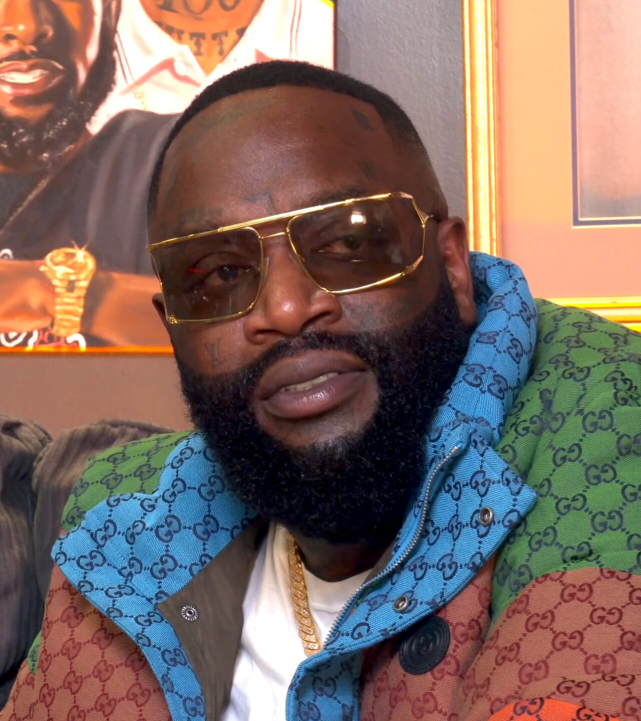 Rick Ross, a dark-skinned man with short black hair, a beard, and gold-framed sunglasses, wearing a colorful jacket with a Gucci pattern