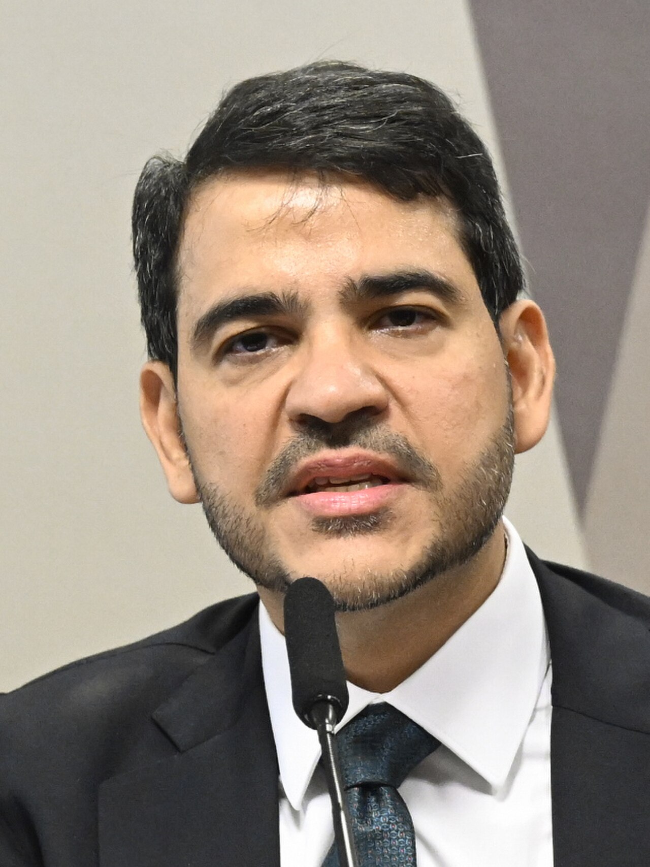 A man with short dark hair, wearing a black suit, white shirt, and dark tie, speaking into a microphone.