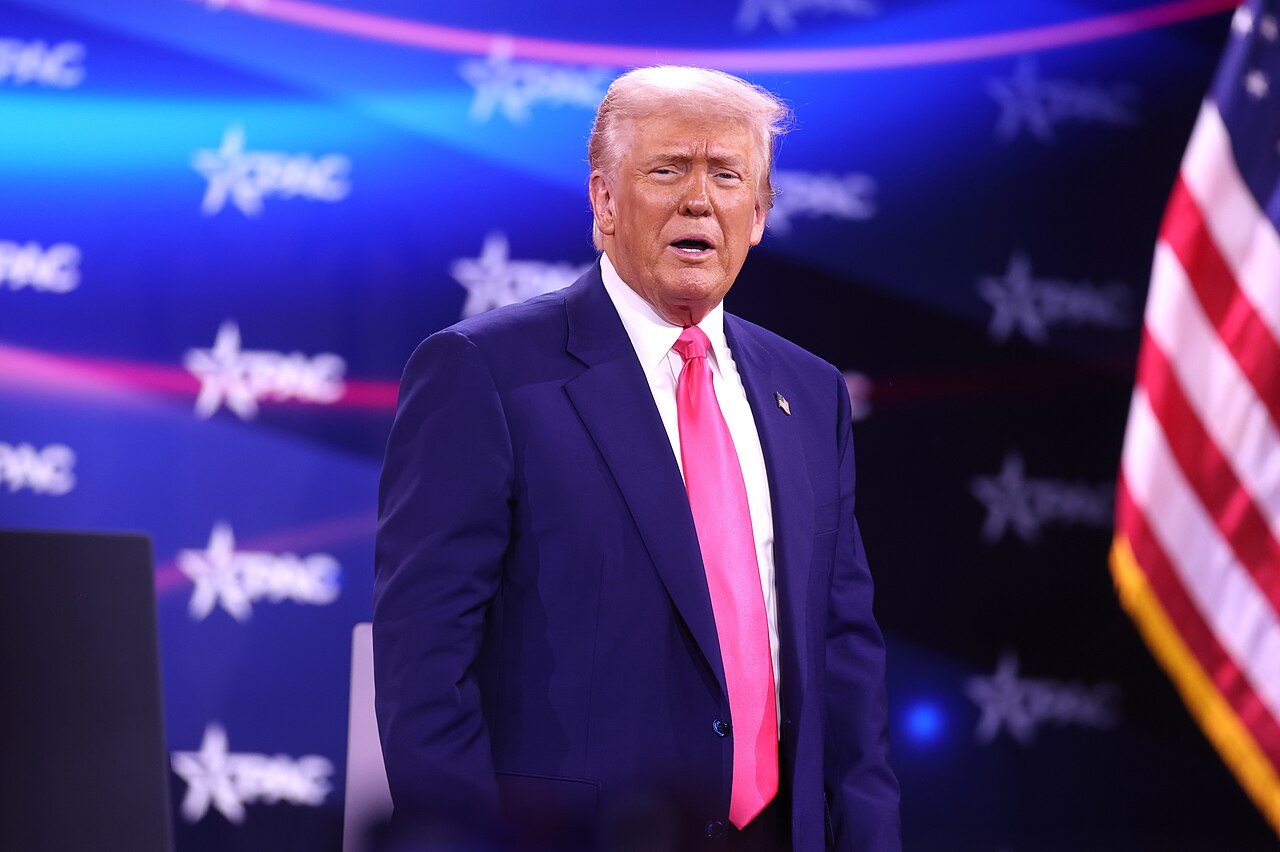Donald Trump in a blue suit and pink tie stands on a stage with a blue background featuring the CPAC logo and an American flag to his right.