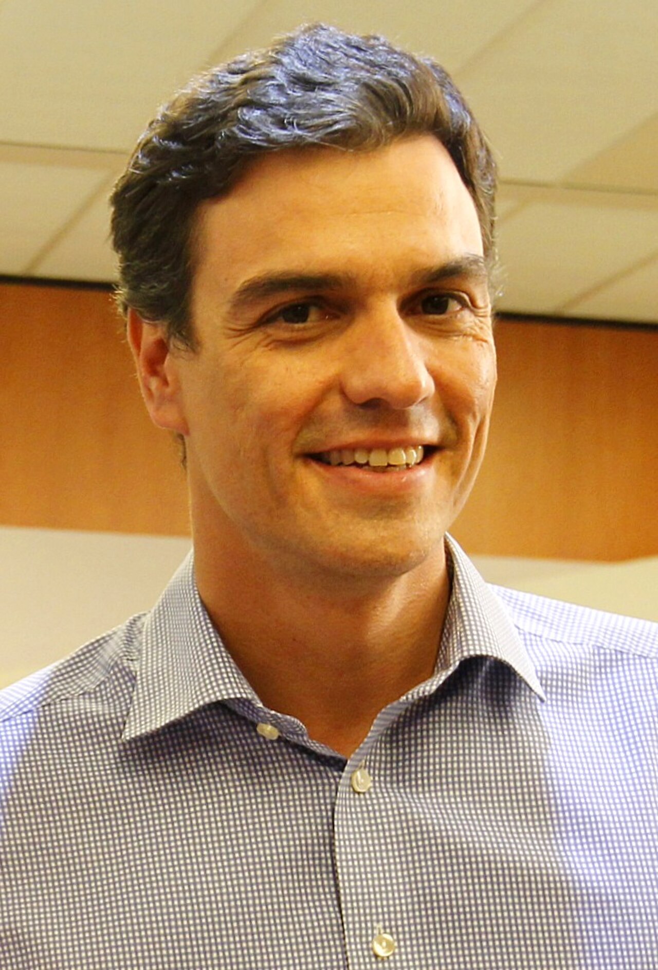A smiling man with short dark hair wearing a blue and white checkered collared shirt