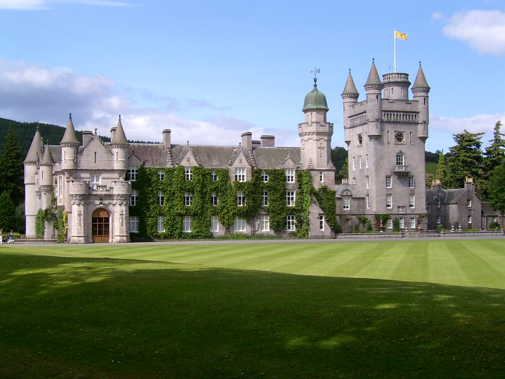 Balmoral Castle with a flag flying from a tower, set against a blue sky with clouds, and a manicured lawn in the foreground.