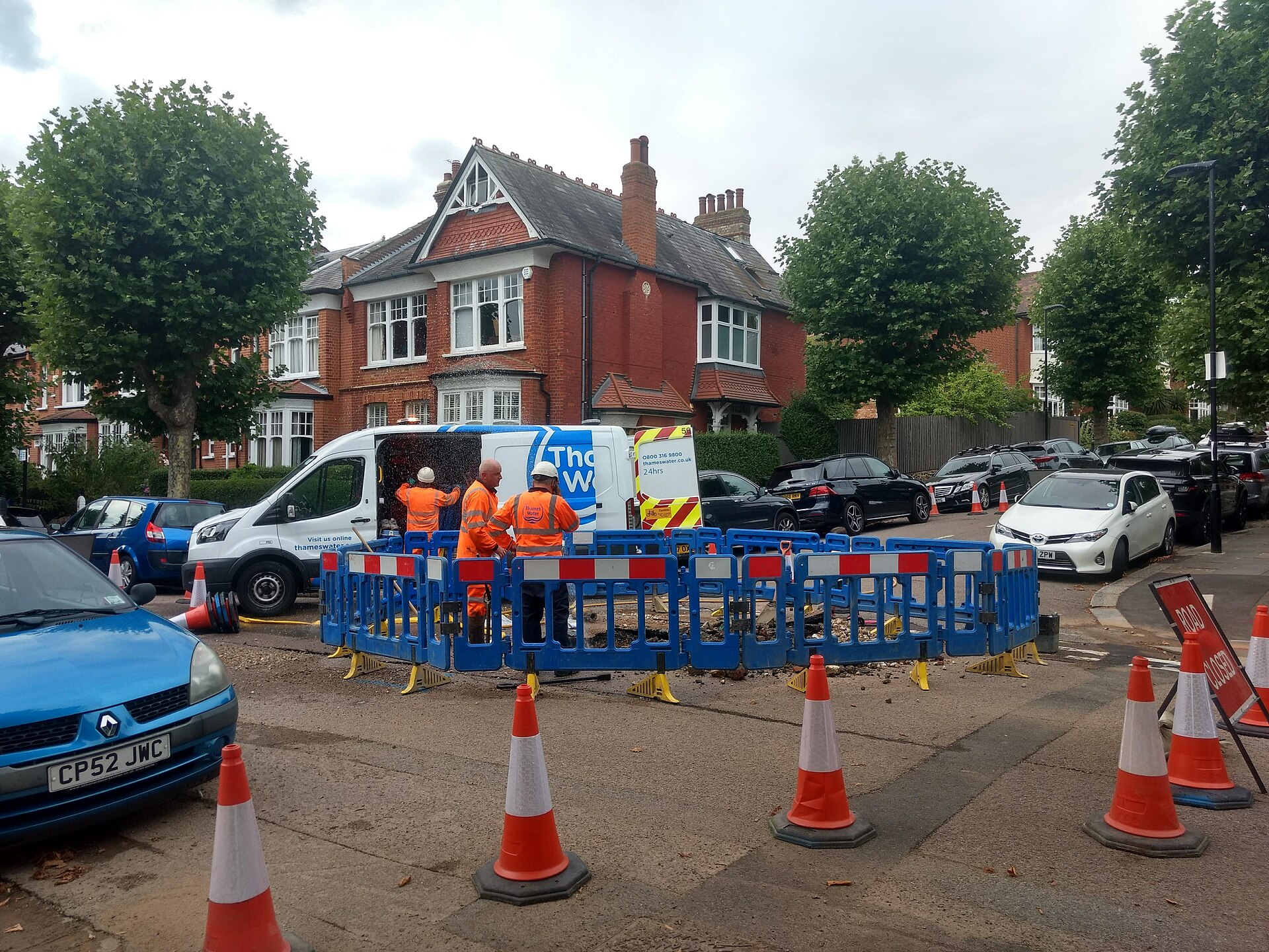 Three workers in orange high-visibility vests stand near a white van with "Thames Water" written on its side, next to a blue construction barrier on a street.