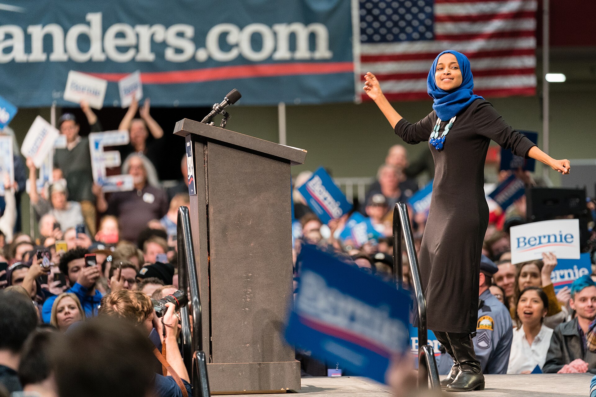 A woman wearing a blue hijab and a long black dress stands on a stage with her arms outstretched, smiling. She is in front of a podium and a crowd of people holding "Bernie" signs. An American flag and a banner that says "Sanders.com" are visible in the background.