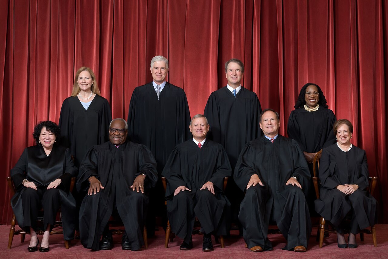 A group of nine justices of the U.S. Supreme Court pose for a formal portrait in front of a red curtain. They are all wearing black robes.