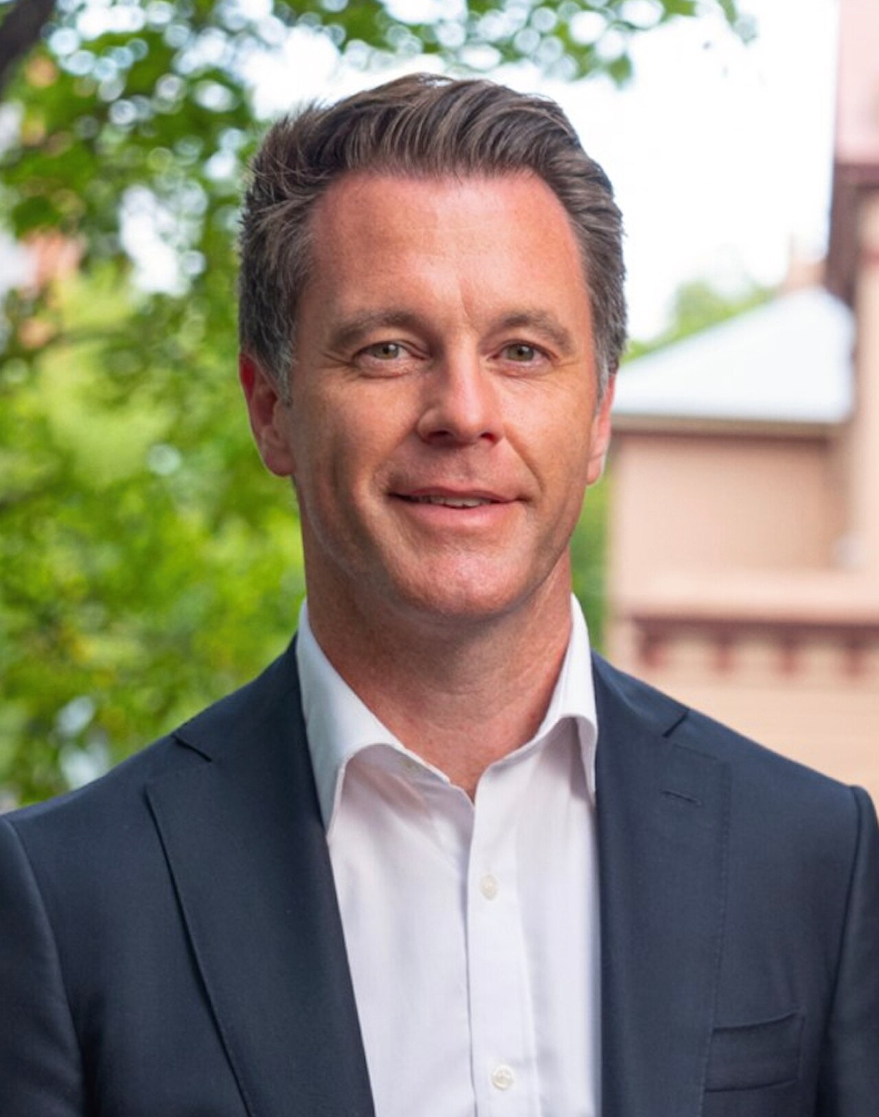 A man with short, graying hair wearing a dark suit jacket and white collared shirt, standing in front of blurred greenery and a building.