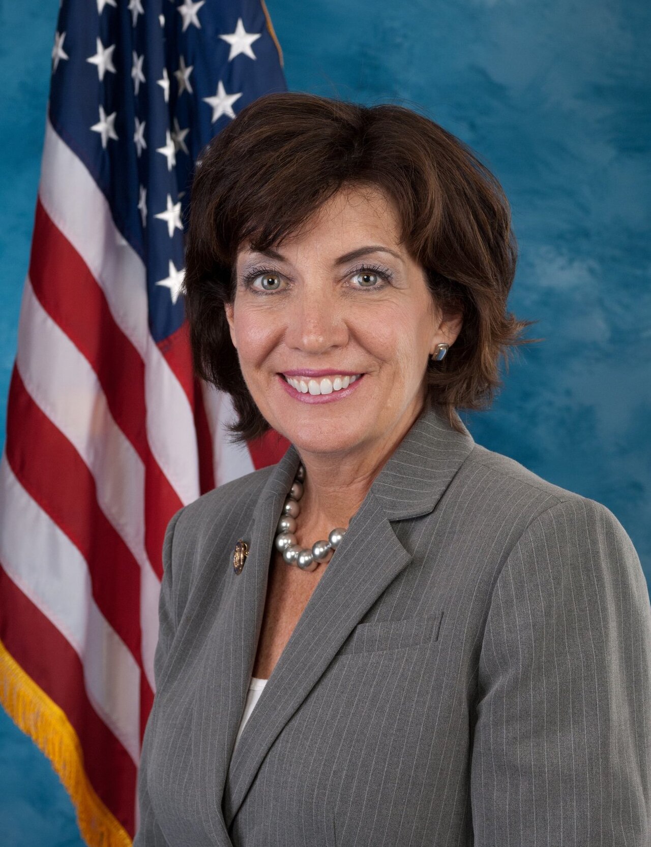 New York Governor Kathy Hochul smiles in a grey pinstripe suit, wearing a pearl necklace, with an American flag behind her.