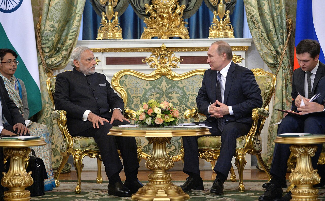 A man with gray hair and beard in a black suit sits in a gold chair, talking to a bald man in a black suit, with a green and white flag and blue and white flag behind them.