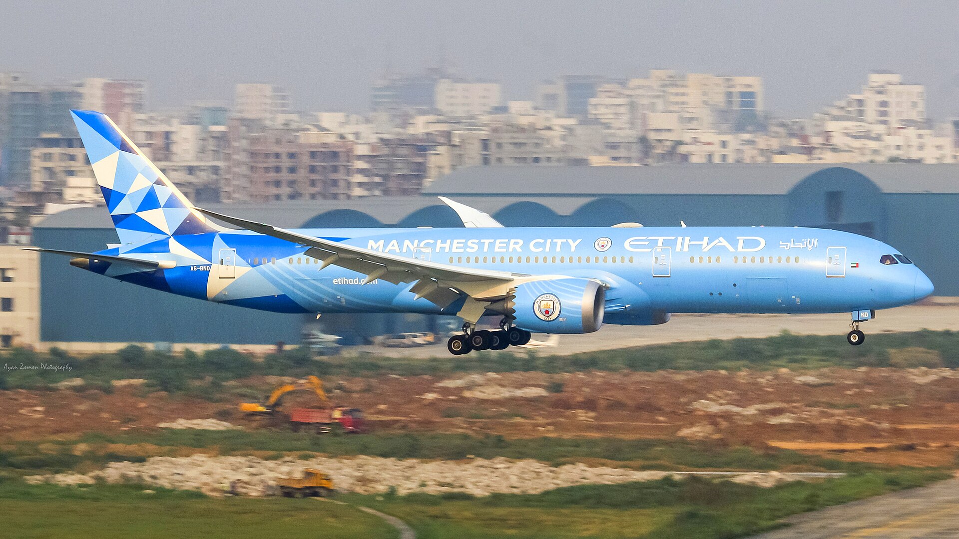 A light blue Etihad Airways airplane with 'MANCHESTER CITY' written on its side is landing. The plane has the Manchester City Football Club crest on its engine and tail.