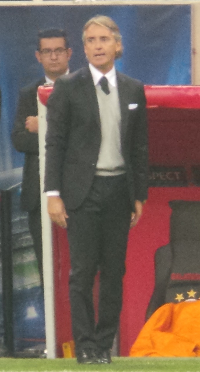 A man with blond hair wearing a black suit, white shirt, black tie, and gray sweater standing on a sports field.