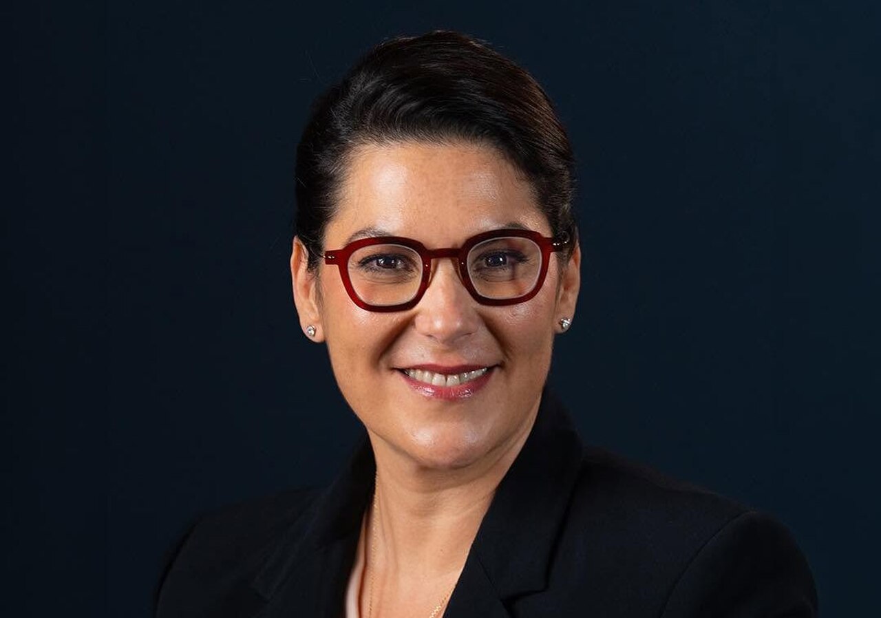A smiling woman with dark hair, red glasses, and a black blazer